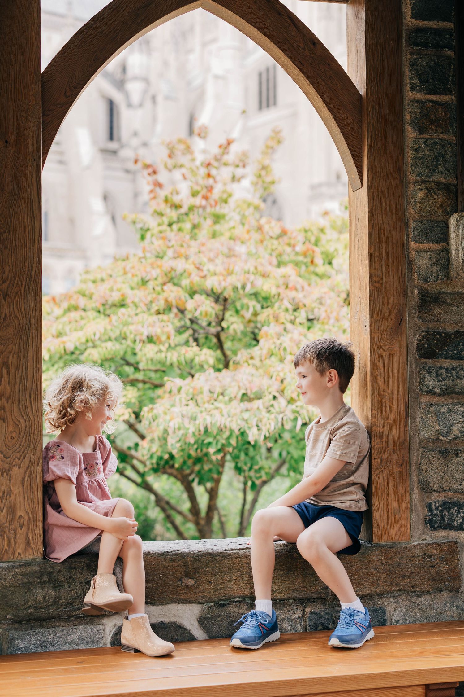 A young girl in a pink dress and a boy in blue sneakers sit on a stone ledge under a wooden arch facing a blooming tree.