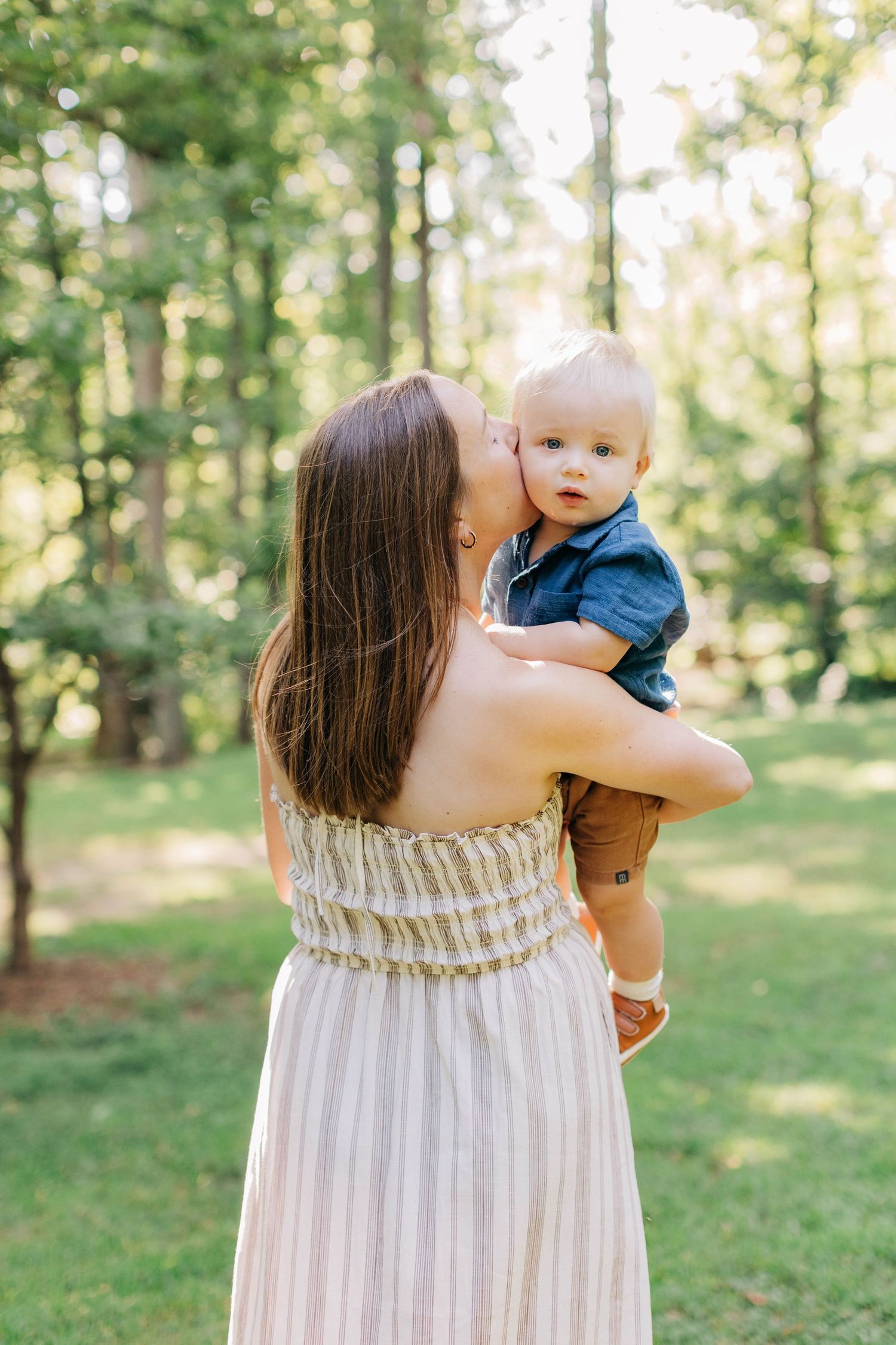 A mother in a striped dress kisses her baby boy outdoors in a lush green park setting.