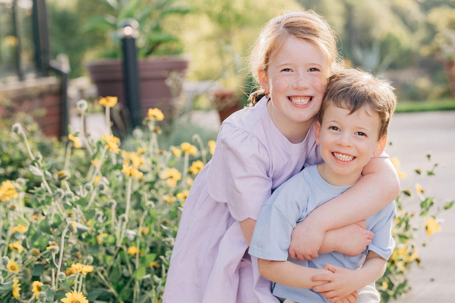 Two smiling children hugging outdoors near yellow flowers, a girl in lavender dress embracing a younger boy in blue shirt.