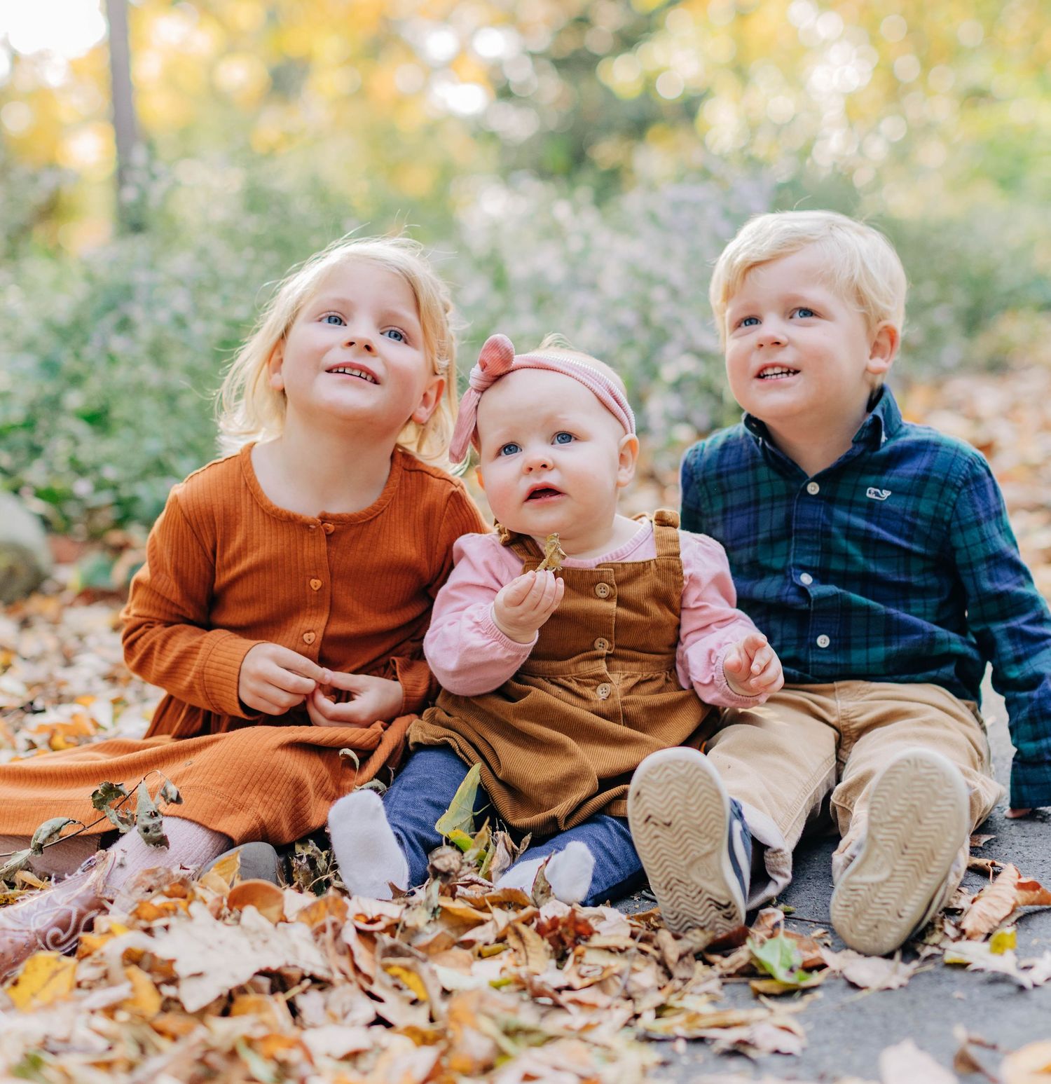 Three young blonde siblings sit together in autumn leaves, two toddlers in fall outfits flanking a smiling baby girl.