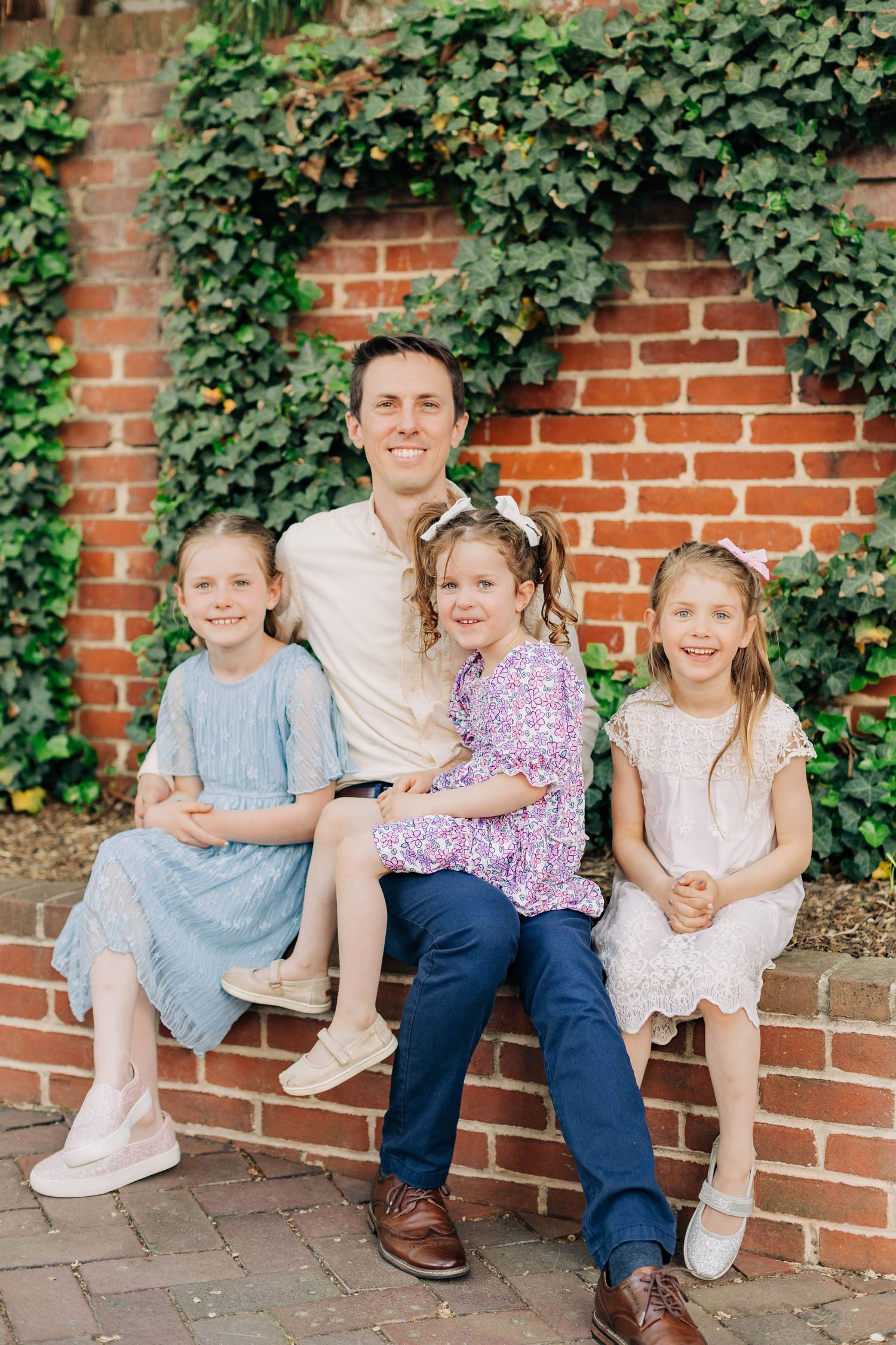 A smiling man sits on a brick wall with three young girls in pastel dresses, posed in front of ivy-covered brick.