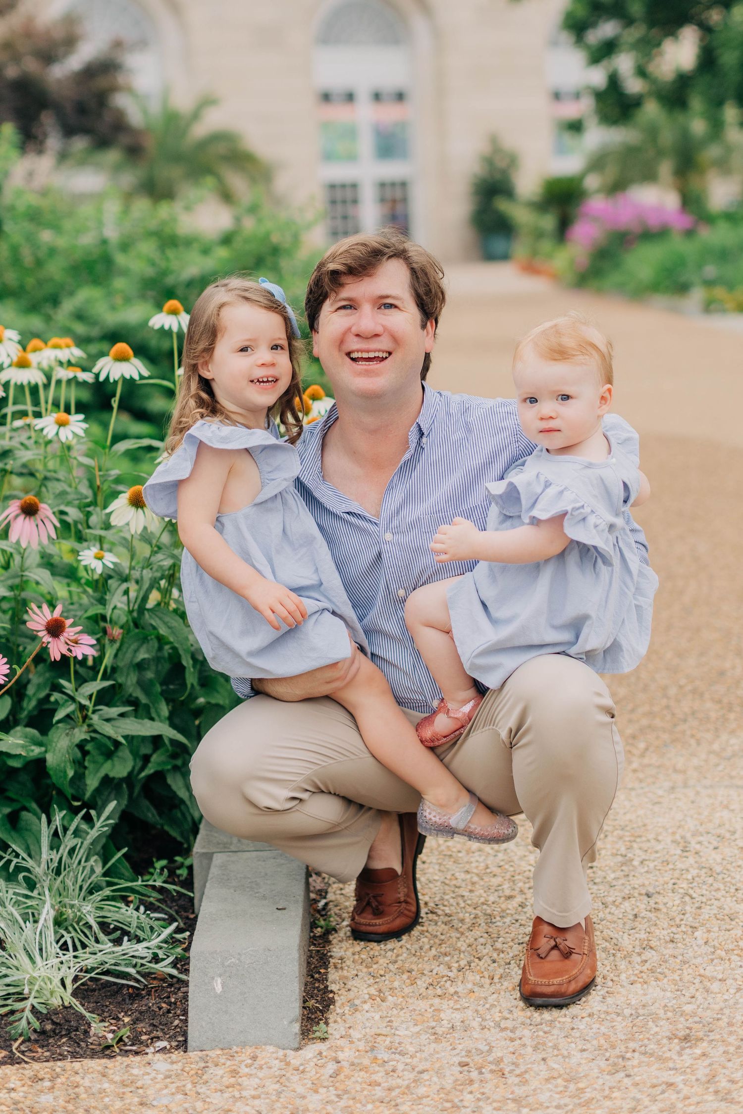 A smiling man crouches in a garden holding two young girls in matching blue dresses near blooming flowers.