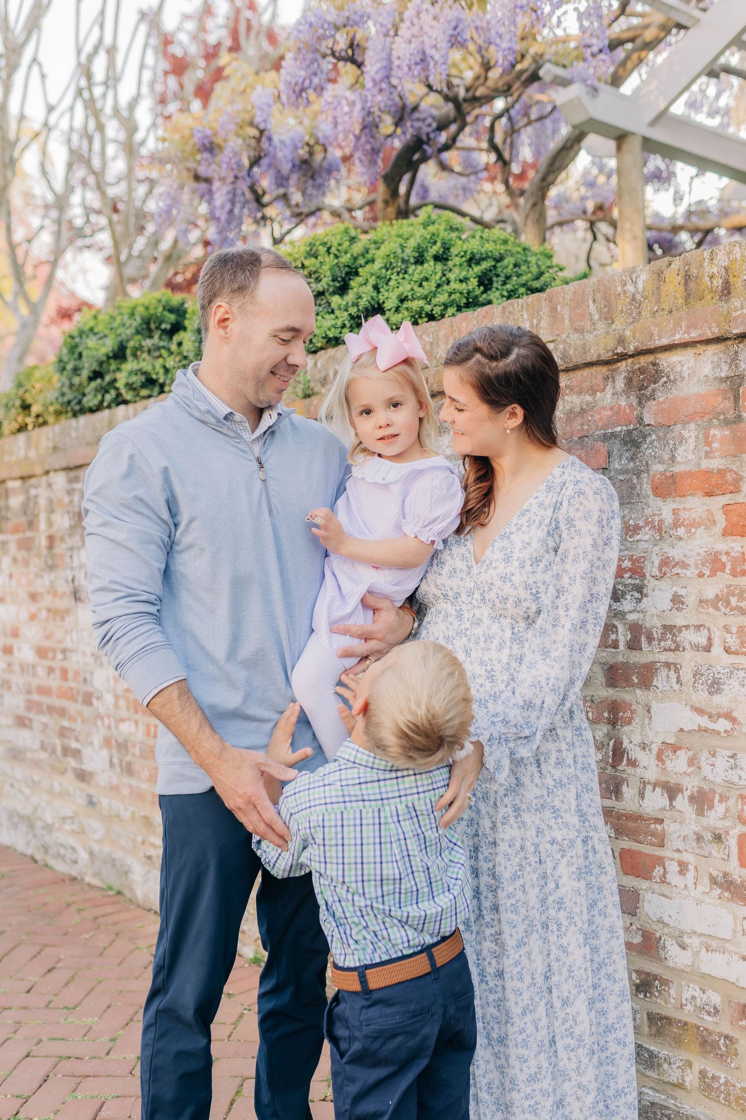 Family of four posing outdoors near brick wall with blooming wisteria, parents smiling at baby girl in pink.