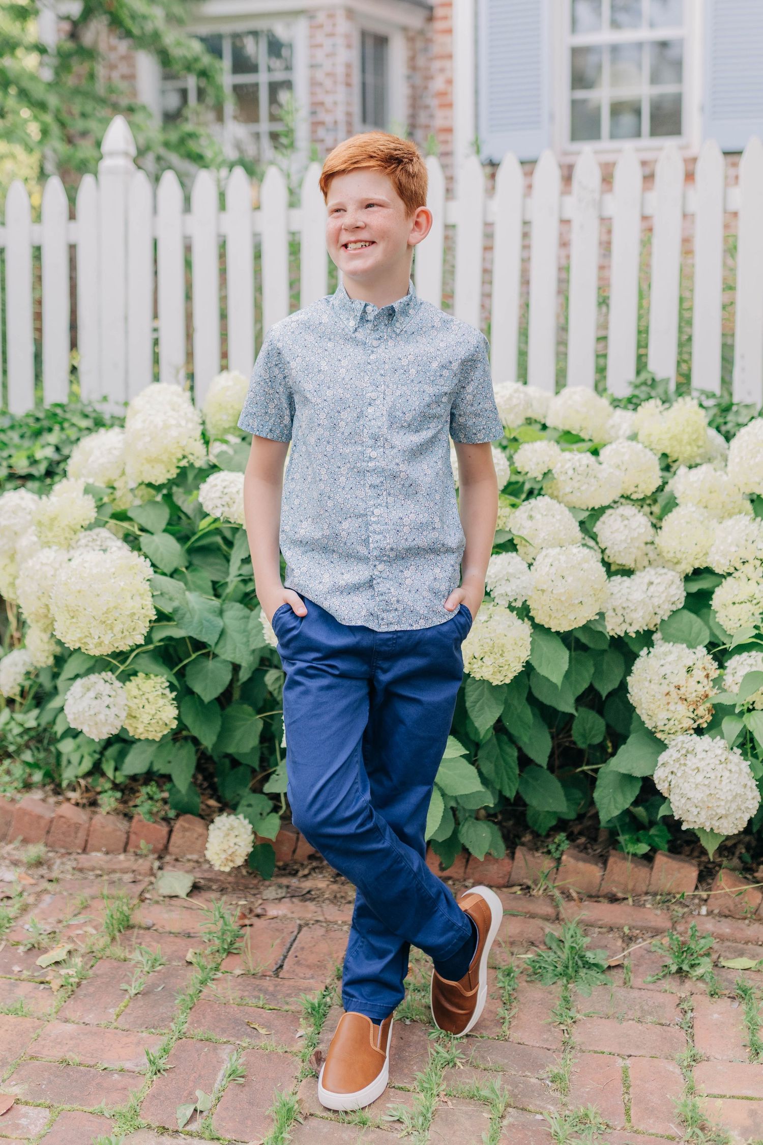 Smiling red-haired boy in blue pants and gray shirt posing outdoors near white hydrangeas and picket fence.