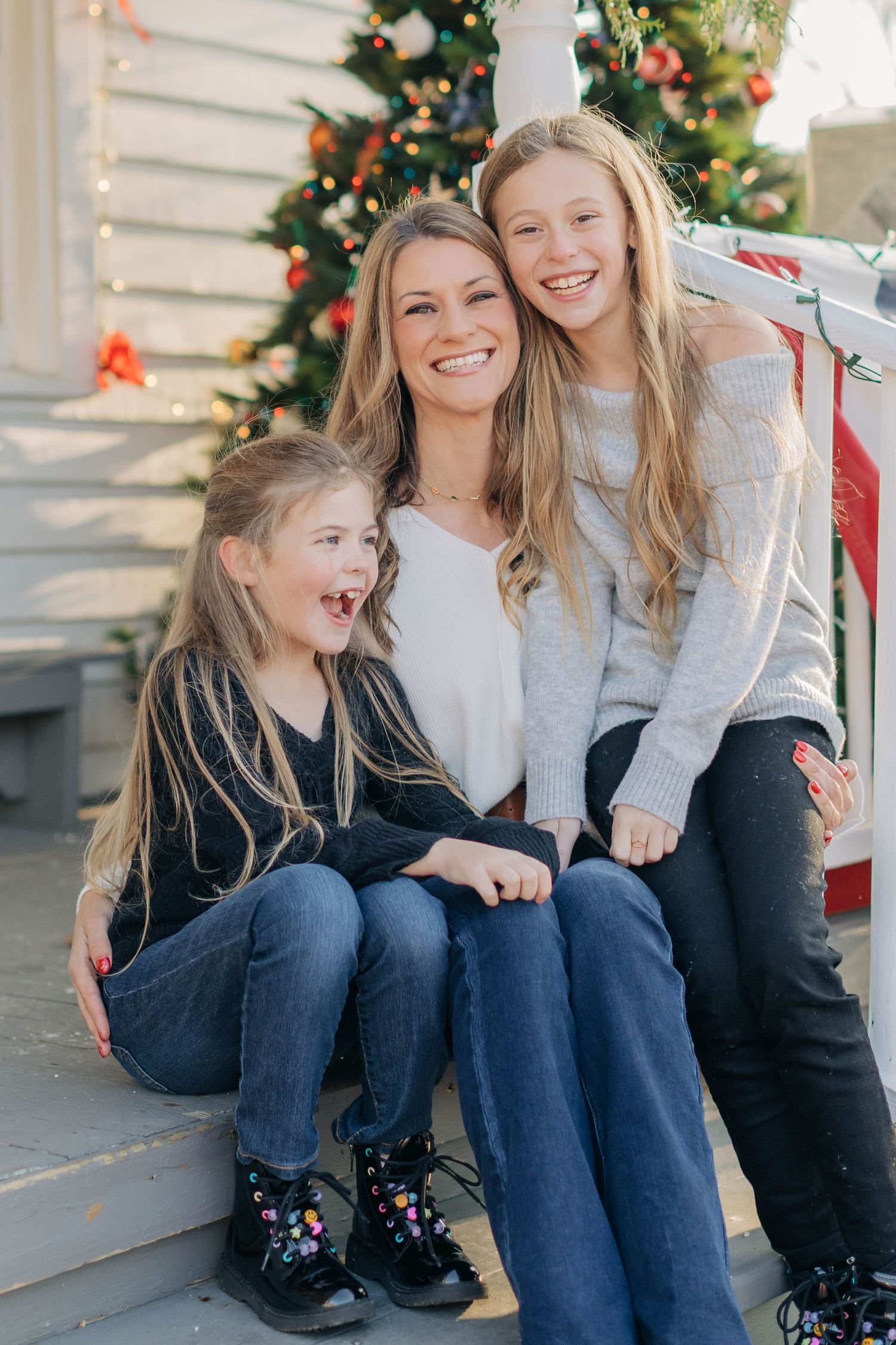 A smiling mother and two daughters pose on porch steps in front of a decorated Christmas tree for a holiday family photo.