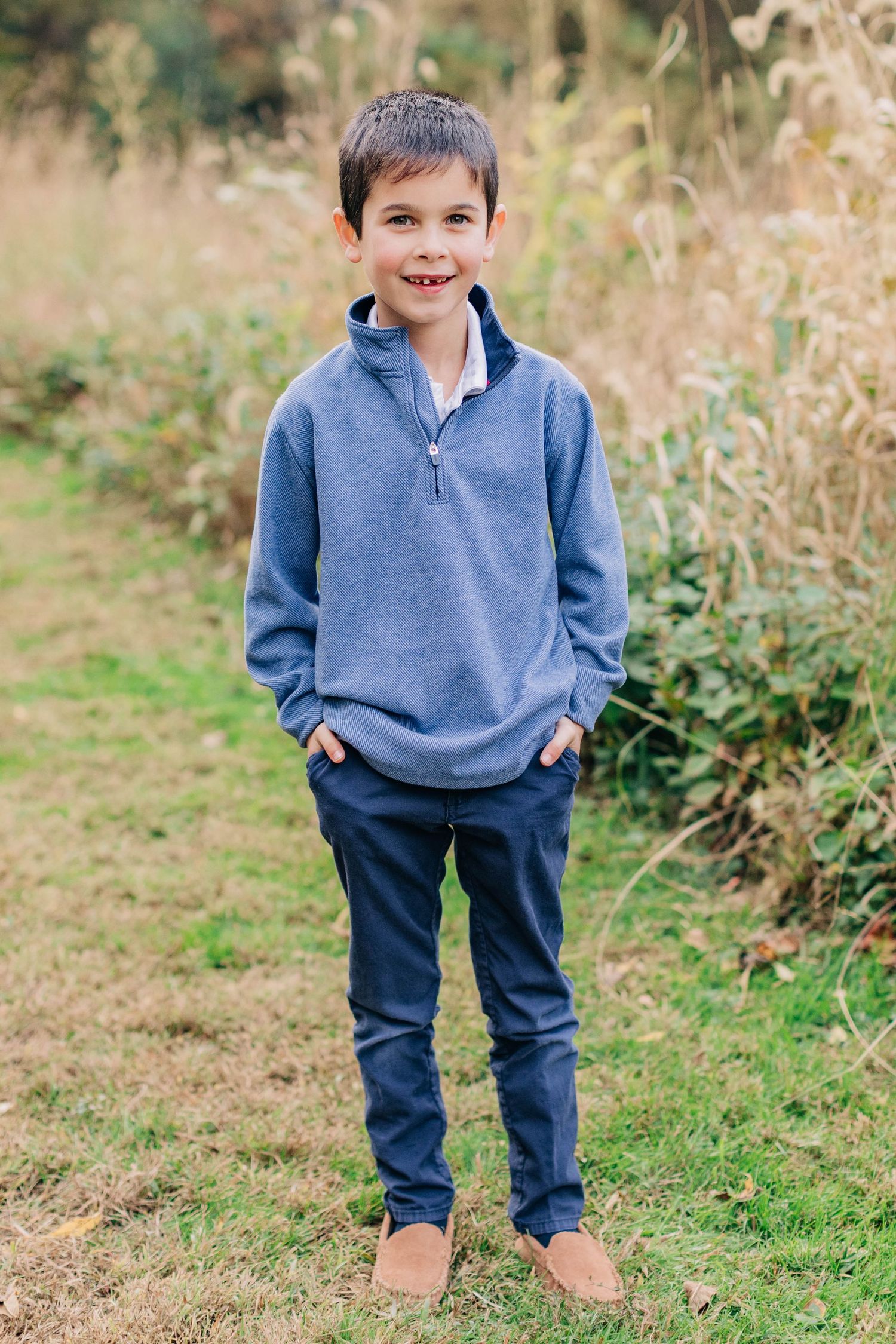 Smiling boy in blue pullover and navy pants stands confidently outdoors with hands in pockets.