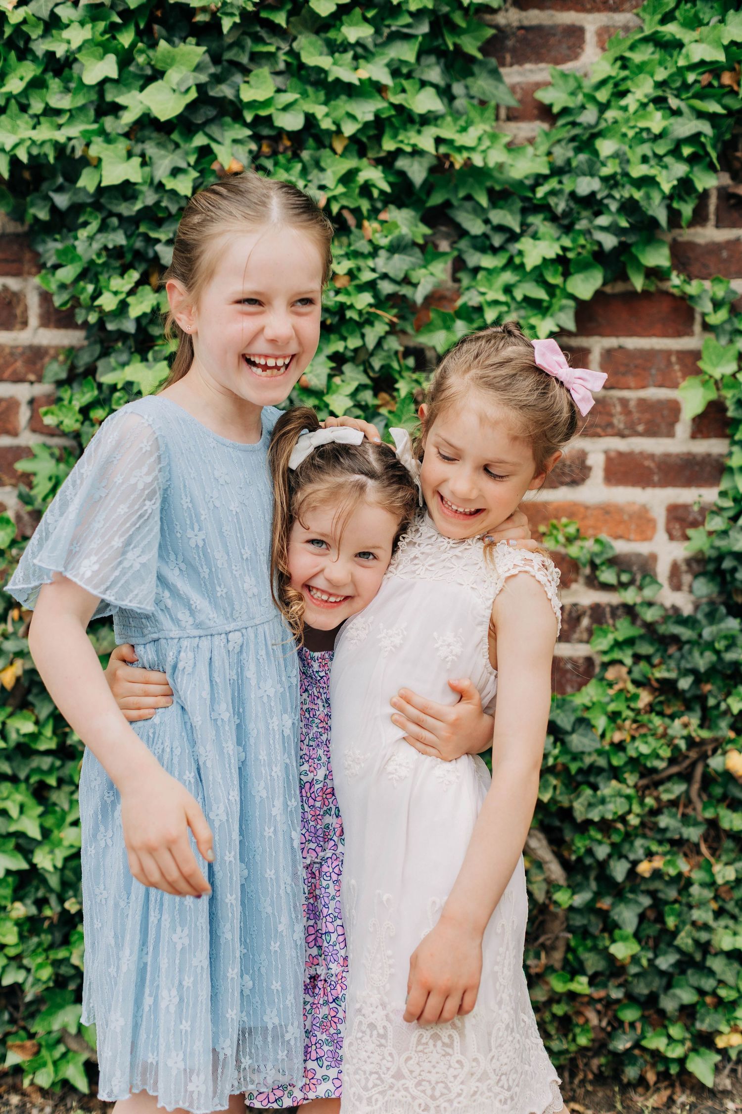 Three young girls in dresses hugging and smiling in front of an ivy-covered brick wall.