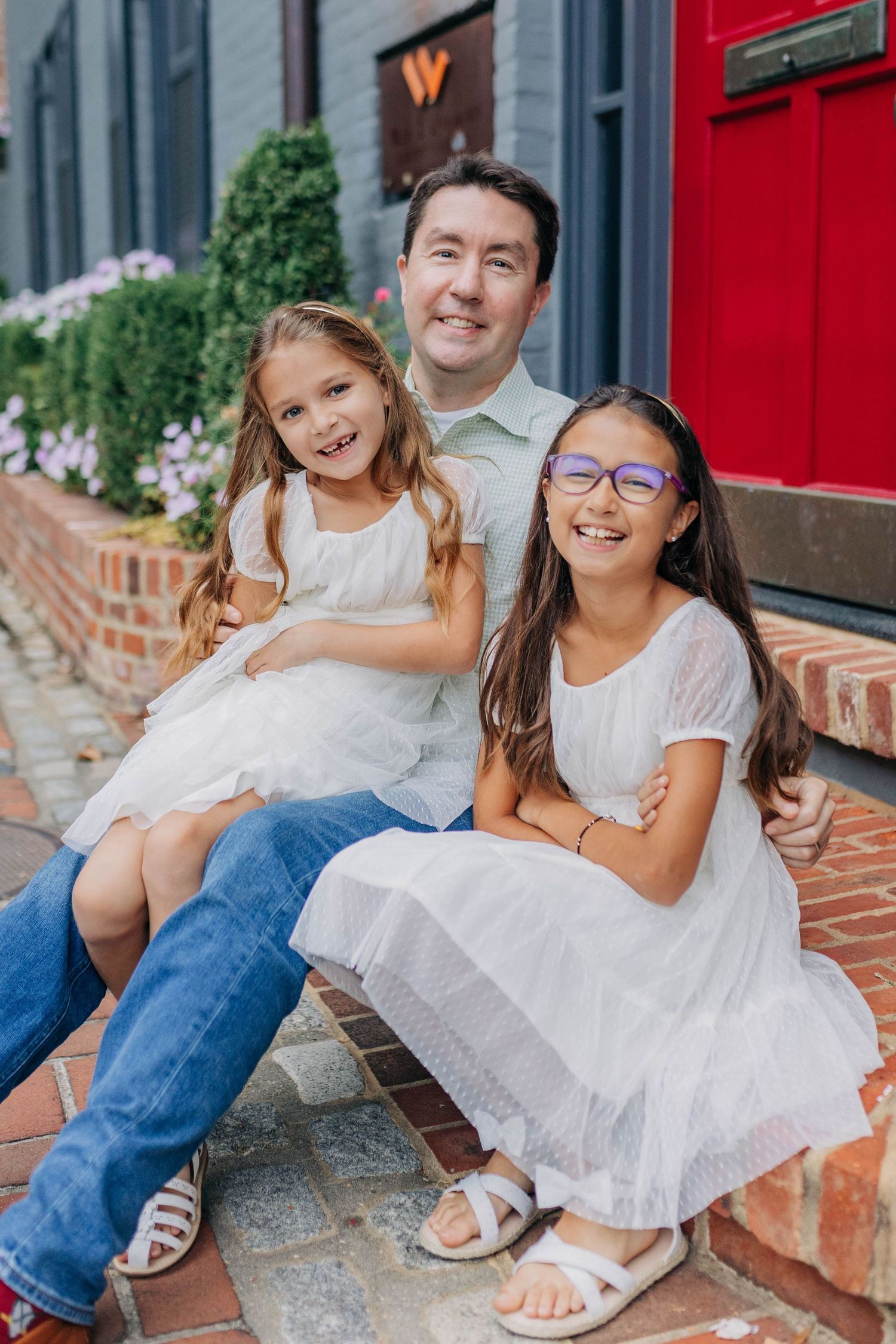 Father sitting on brick steps with two young daughters in white dresses, smiling.