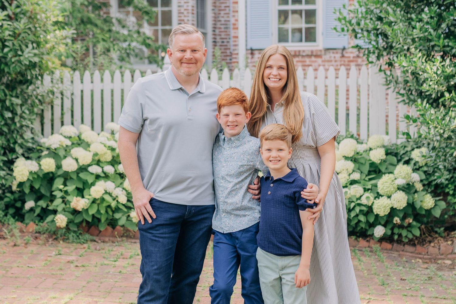 Family of four smiling outdoors near white fence and hydrangeas, parents with two red-haired boys in preppy attire.