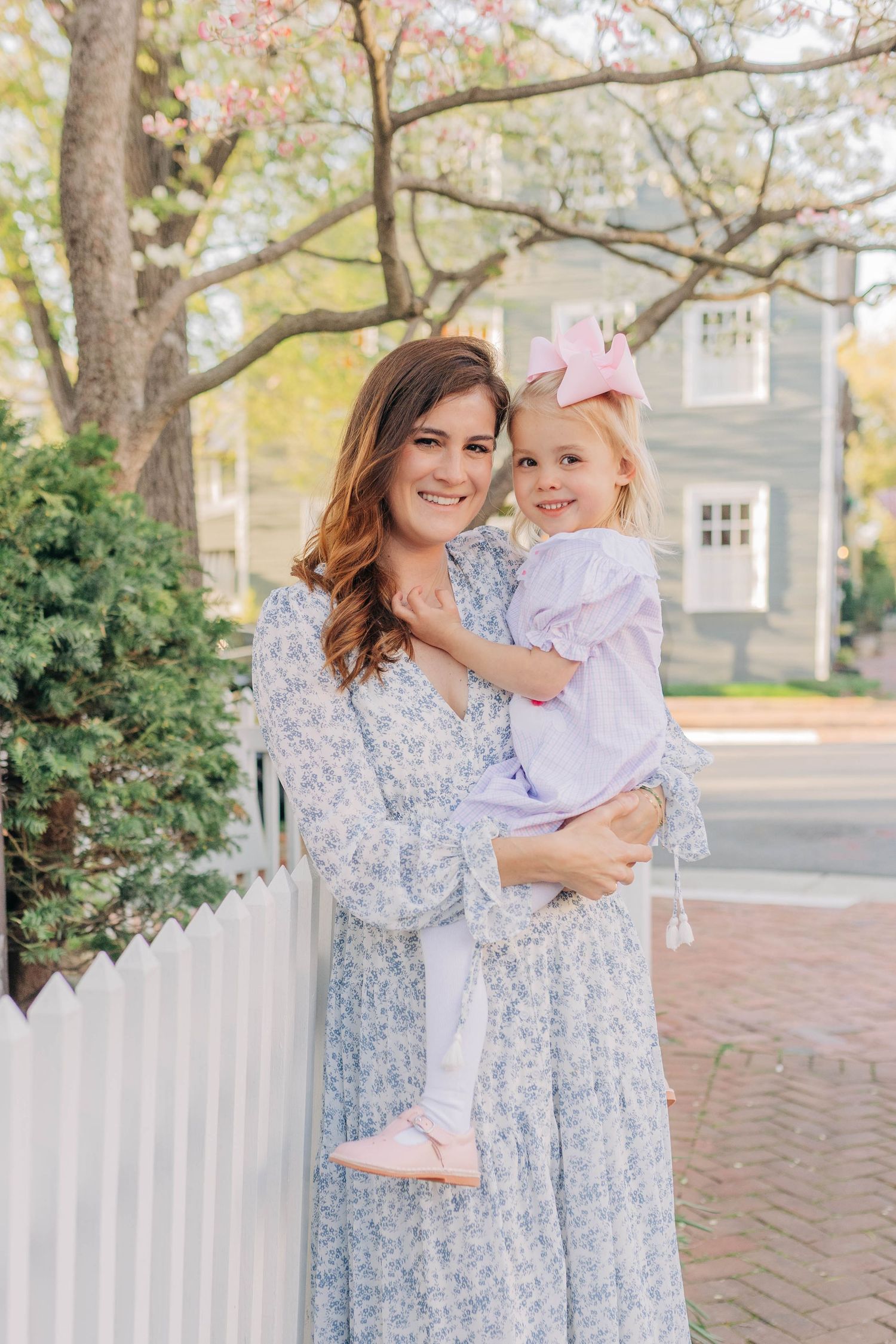 Woman in floral dress holding young girl in lavender outfit with pink bow outdoors near white fence.