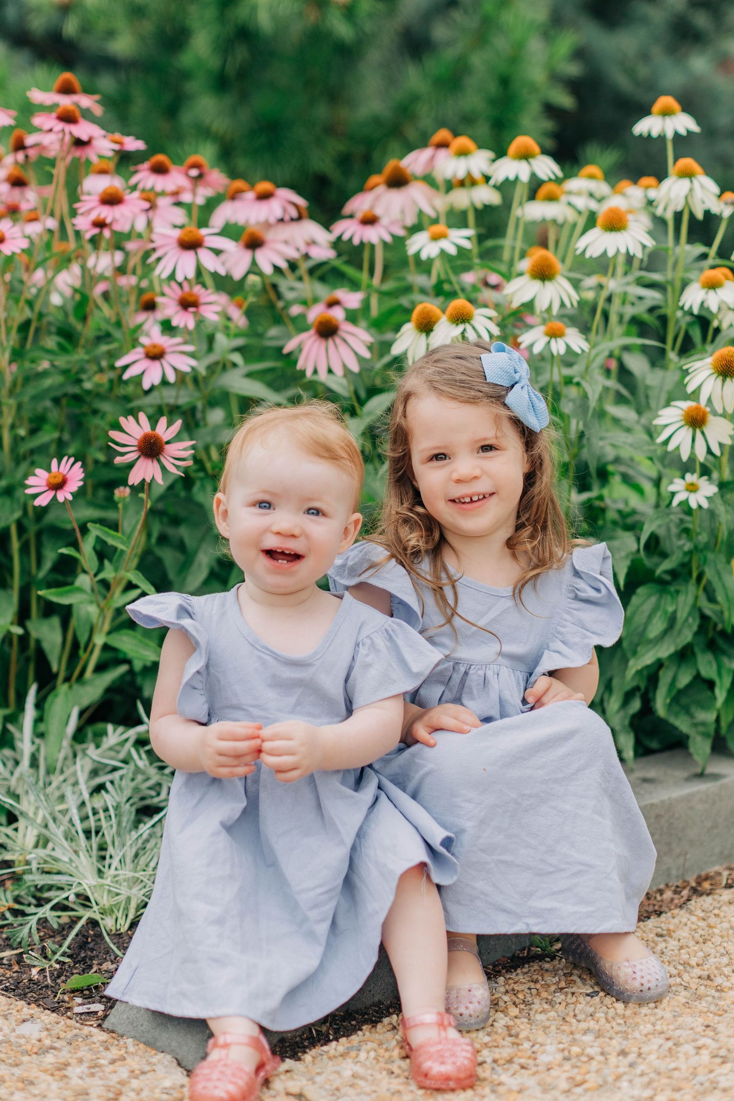 Two young girls in matching blue dresses smile together in front of pink echinacea flowers in a garden.