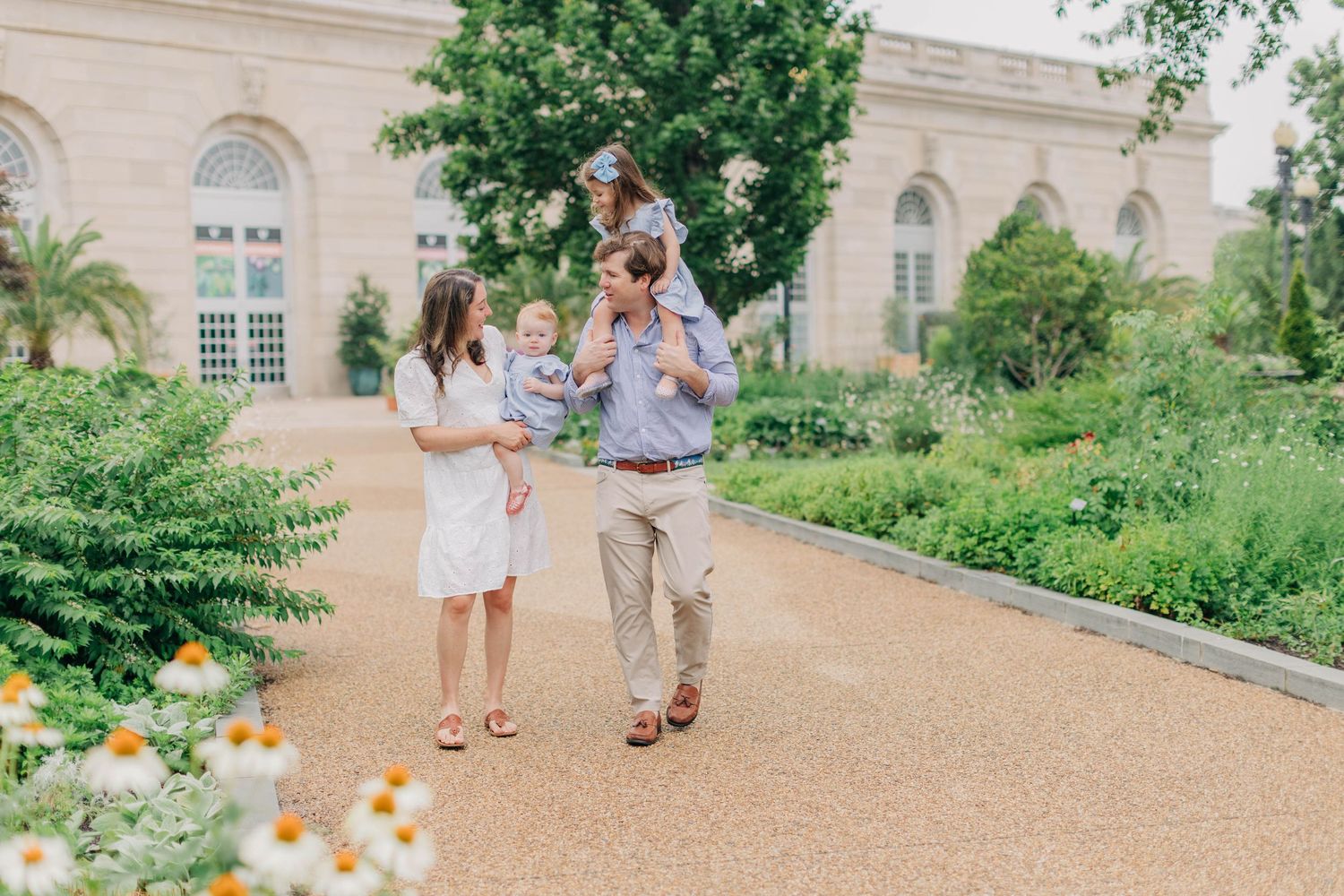 A happy family strolls through a lush garden path, with dad carrying a child on his shoulders near a classic building.
