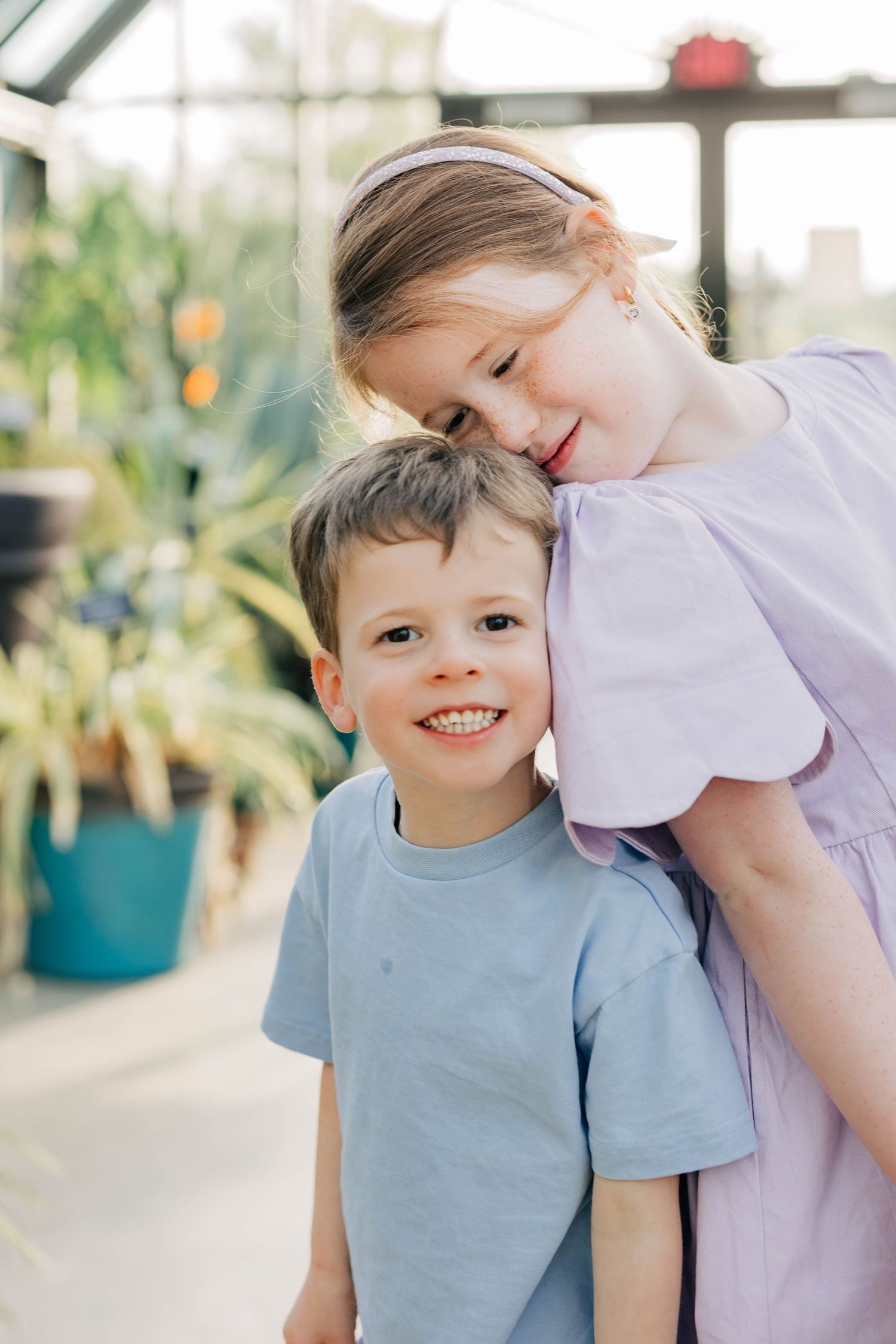 A smiling boy in a blue t-shirt hugged by a girl in a purple dress inside a lush greenhouse.