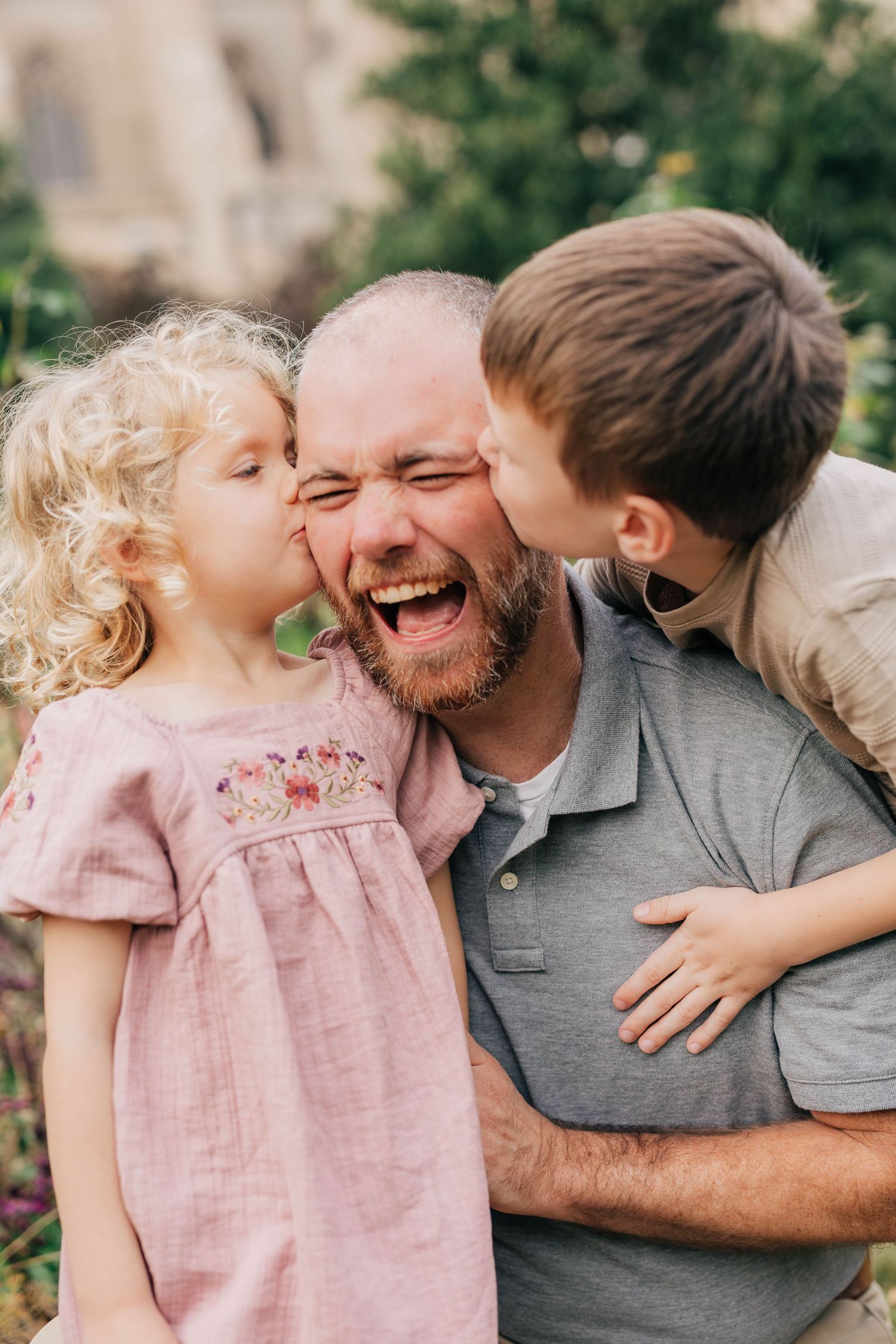 Two laughing children kiss a smiling bearded man on both cheeks in a joyful outdoor family portrait surrounded by greenery.