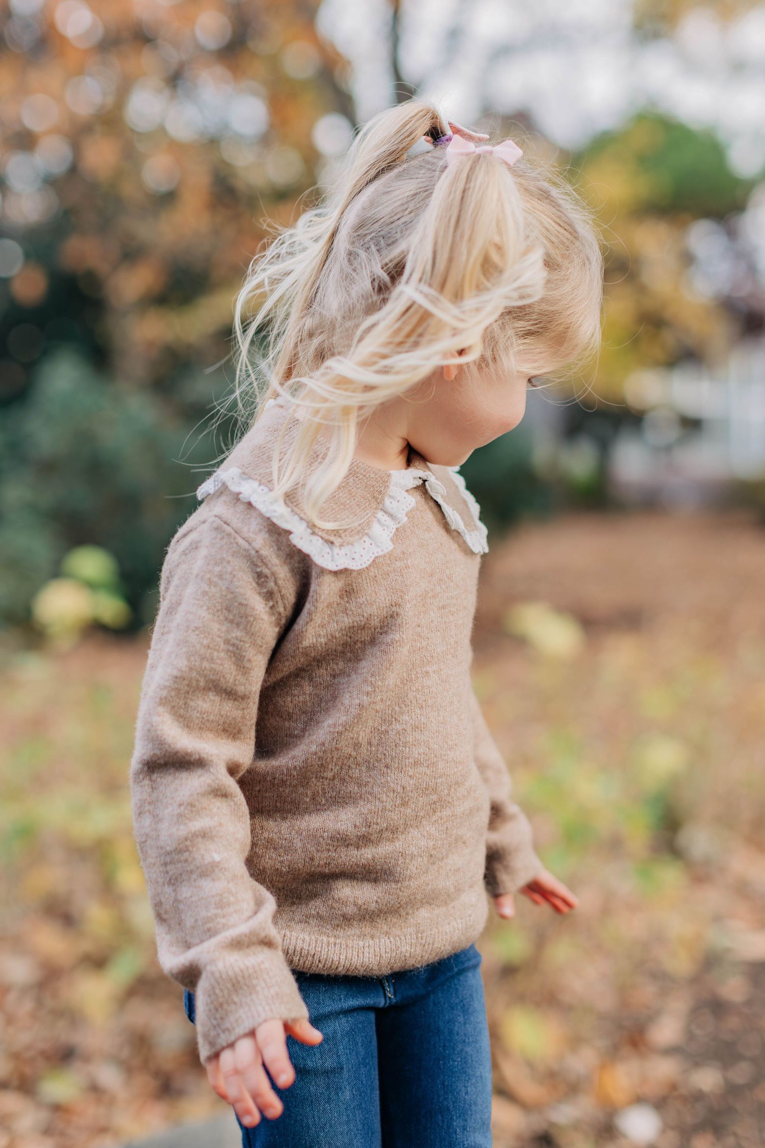 Young blonde girl in a tan sweater playing outdoors in fall leaves.