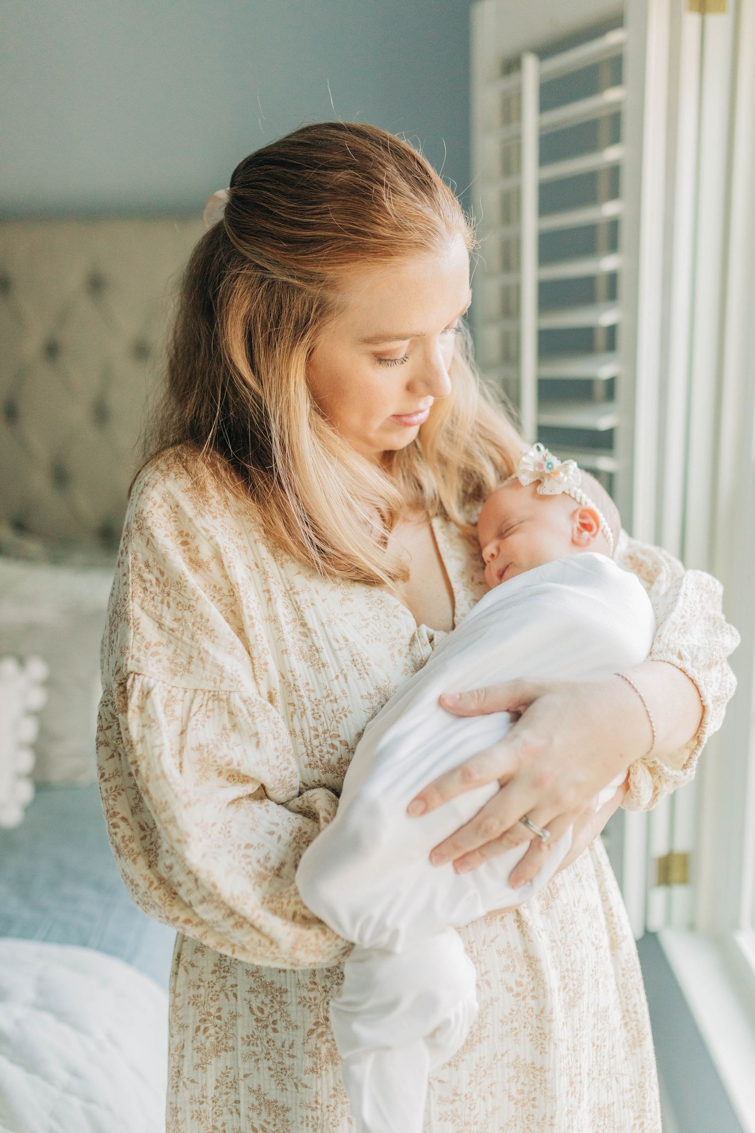 Intimate lifestyle photos of mother in cream dress holding newborn baby in bright nursery.