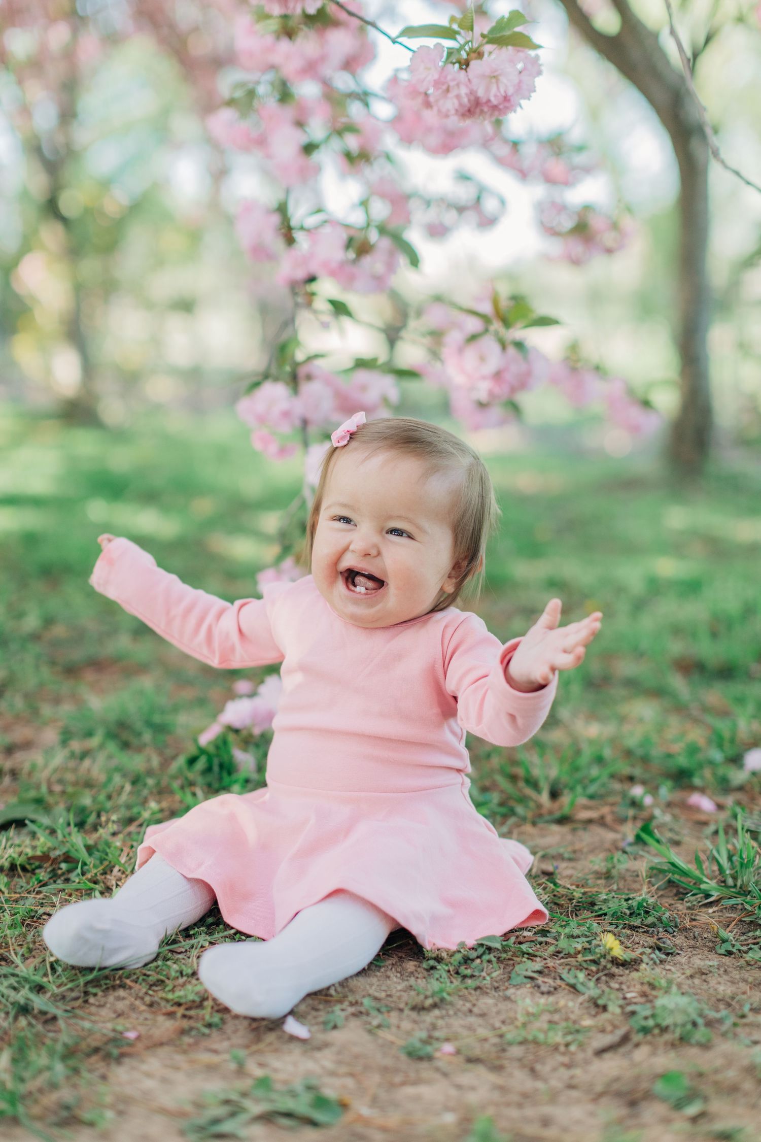 Baby wearing pink outfit laughing while sitting under spring blossoms in garden setting.