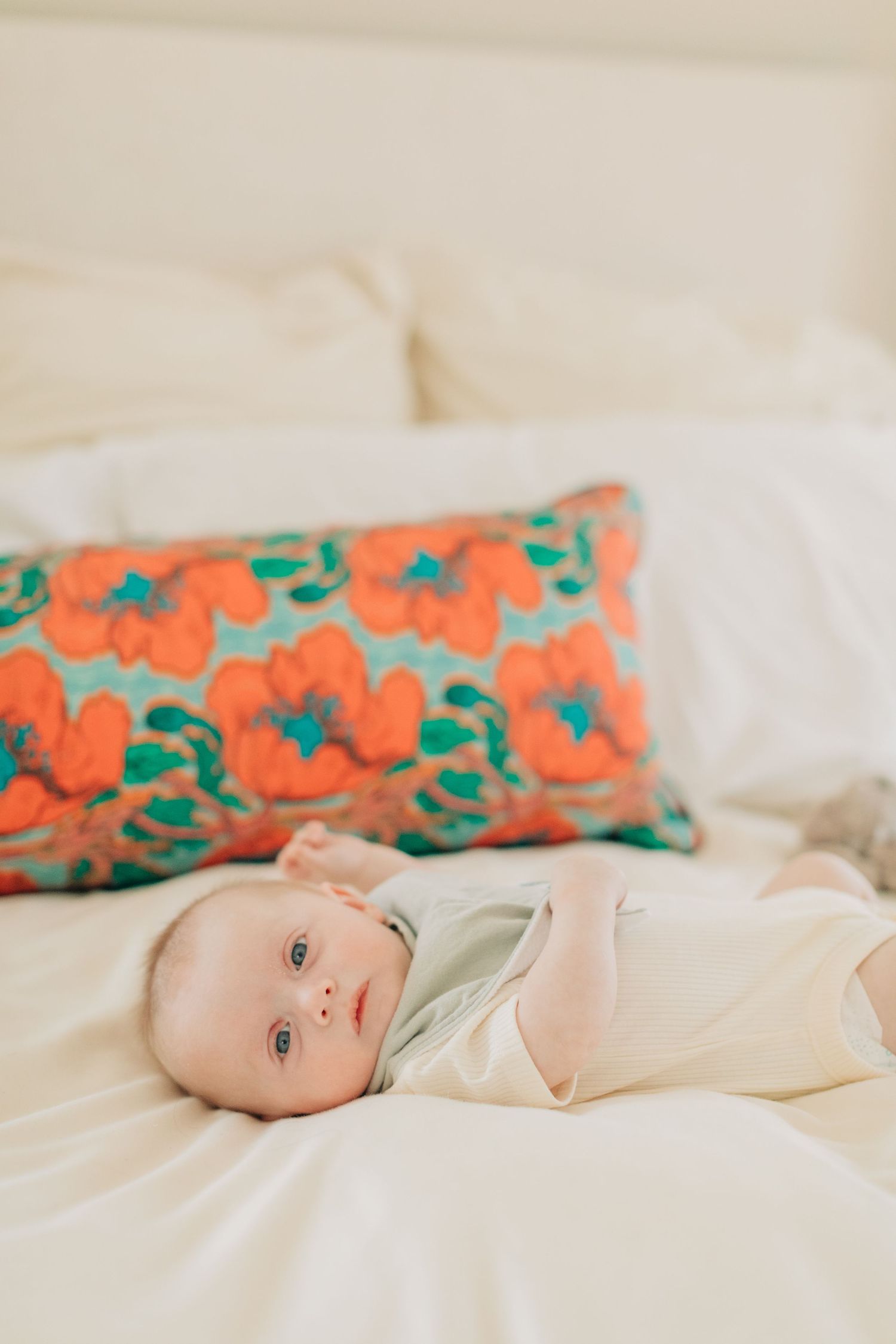Baby lies on white bedding next to colorful orange and teal floral pillow.