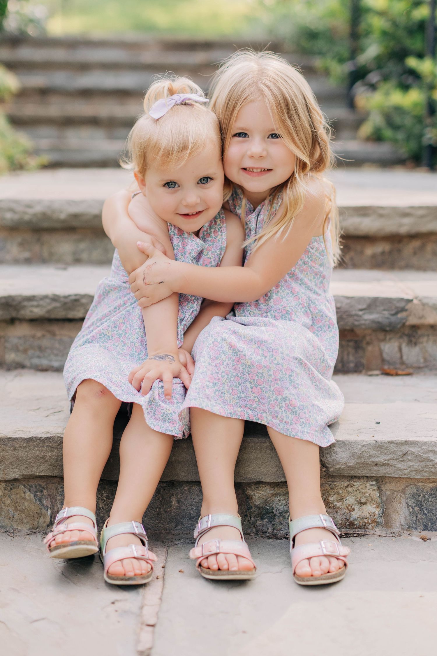 Two young sisters in matching floral dresses hugging on stone steps outdoors.