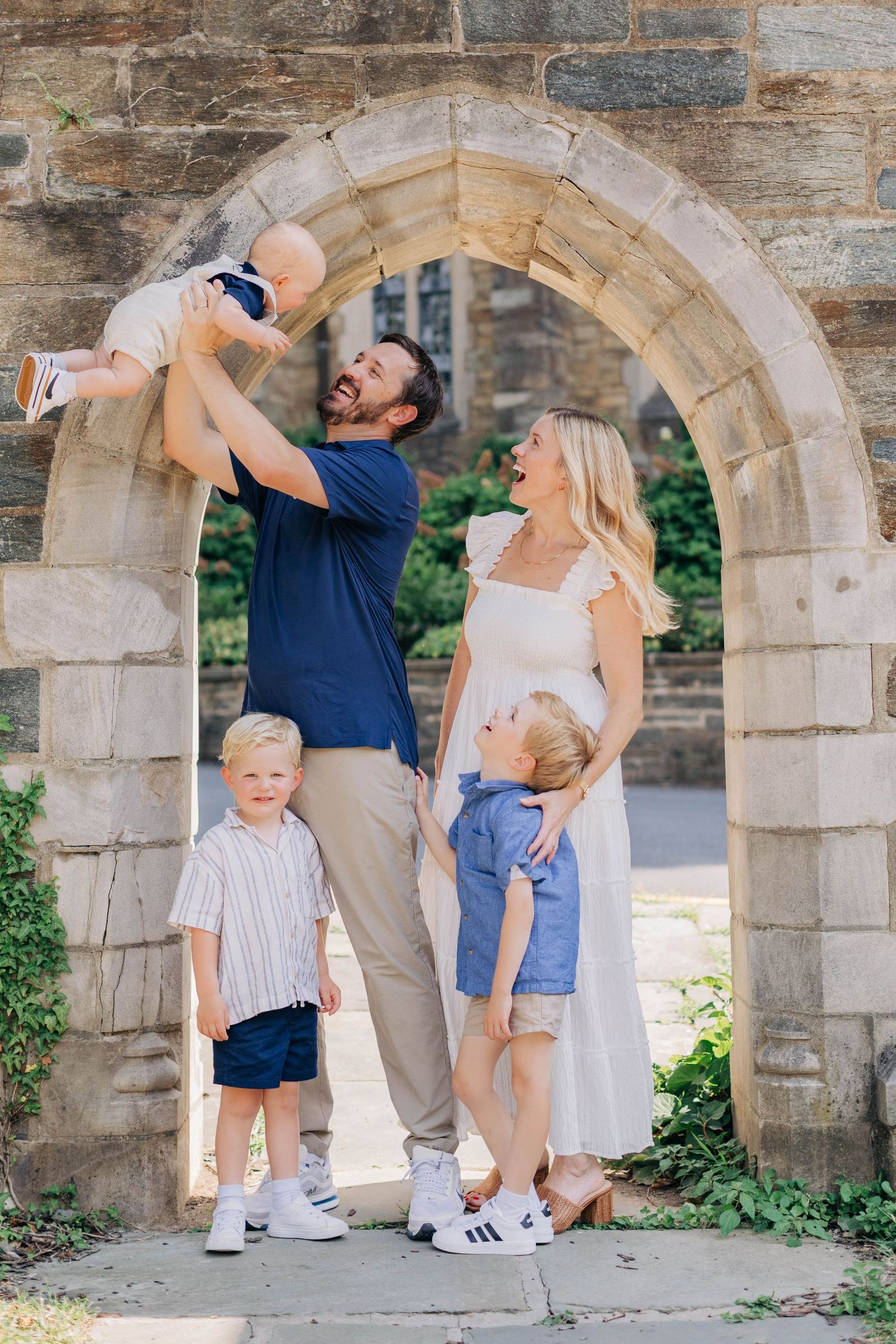 Happy family of five posing under a stone arch outdoors, father lifting baby while two young boys stand nearby.