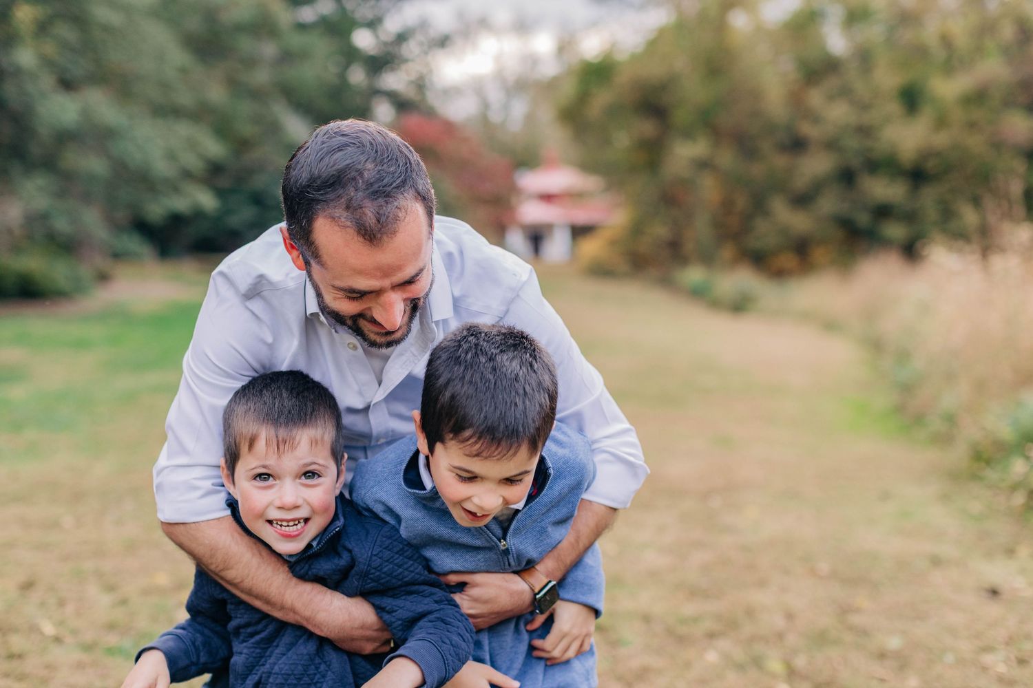 Smiling father playfully hugging two young boys outdoors in a grassy park setting.