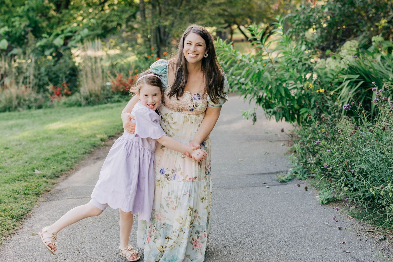 A woman in a floral maxi dress poses with a young girl in a lavender dress on a garden path.