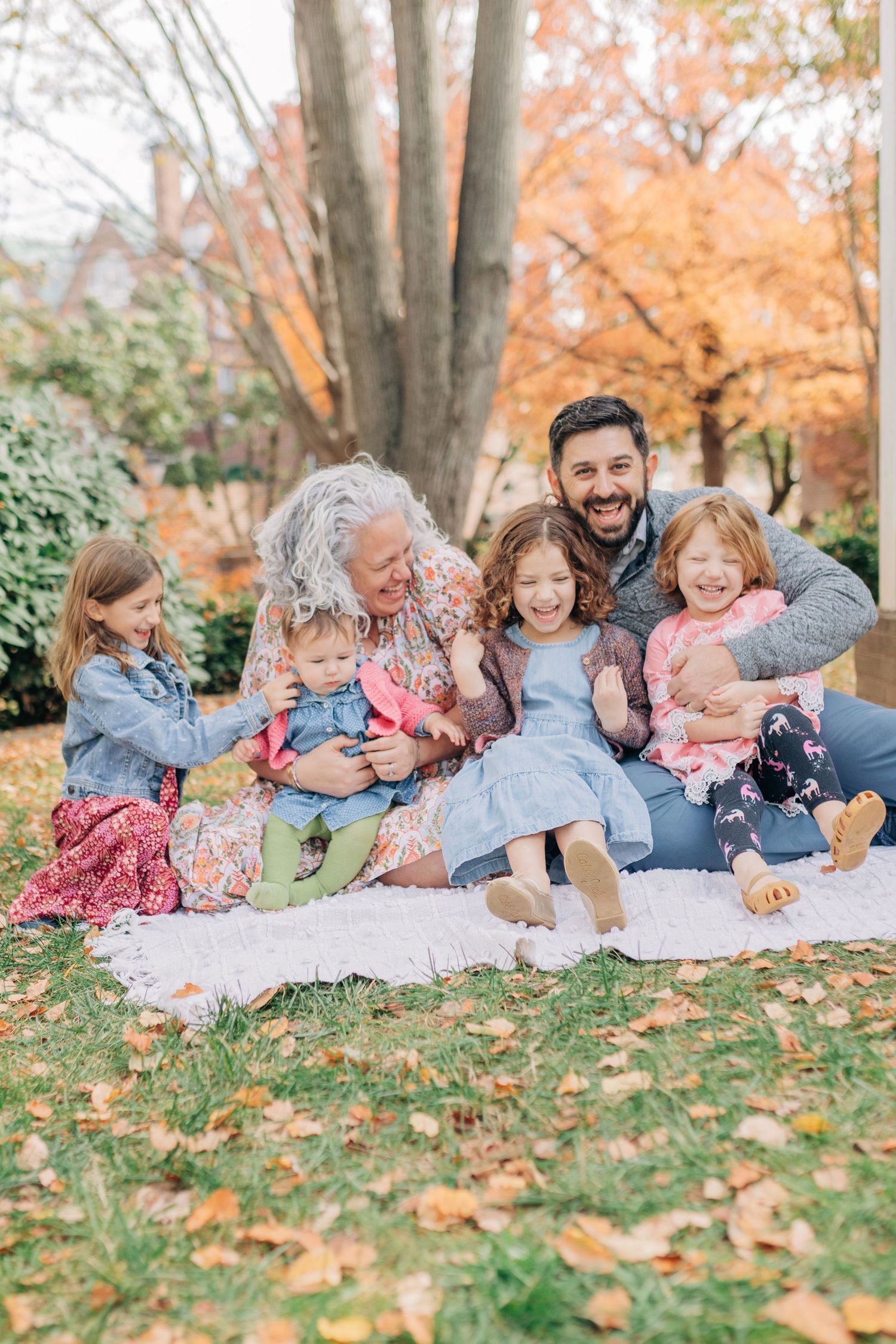 A joyful multigenerational family sits together on a blanket surrounded by fall foliage in a park.