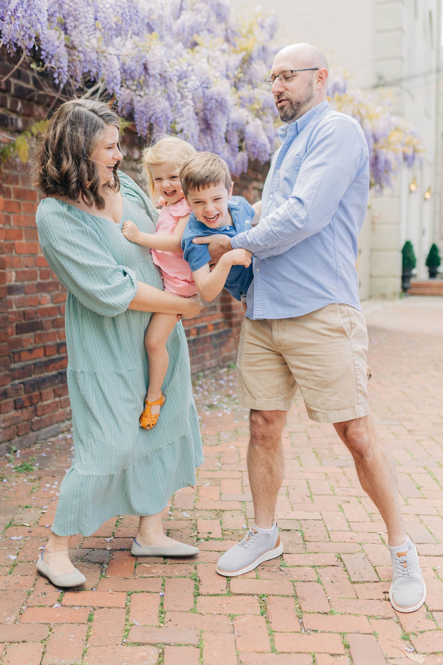 Family of four laughing together under purple wisteria blooms on a brick pathway in spring.