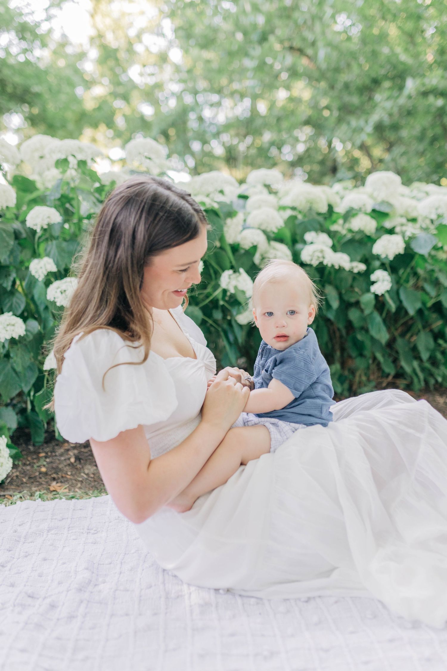 Mother and baby lying on white blanket in garden setting with white flowers.