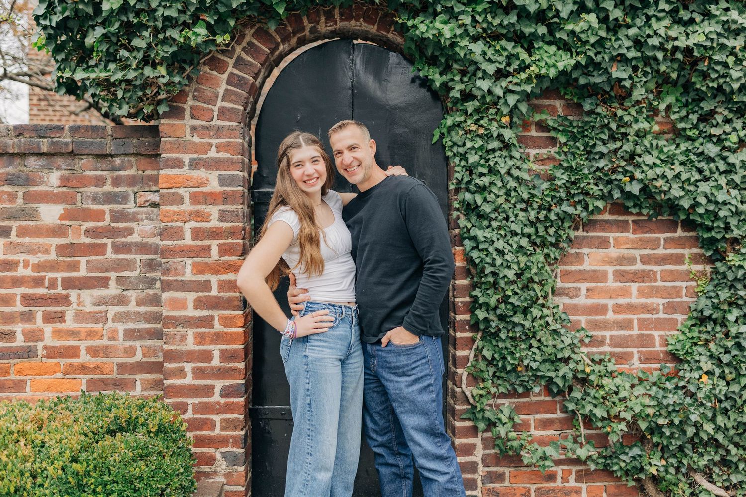 A couple posing together in front of an ivy-covered brick wall with a dark door.