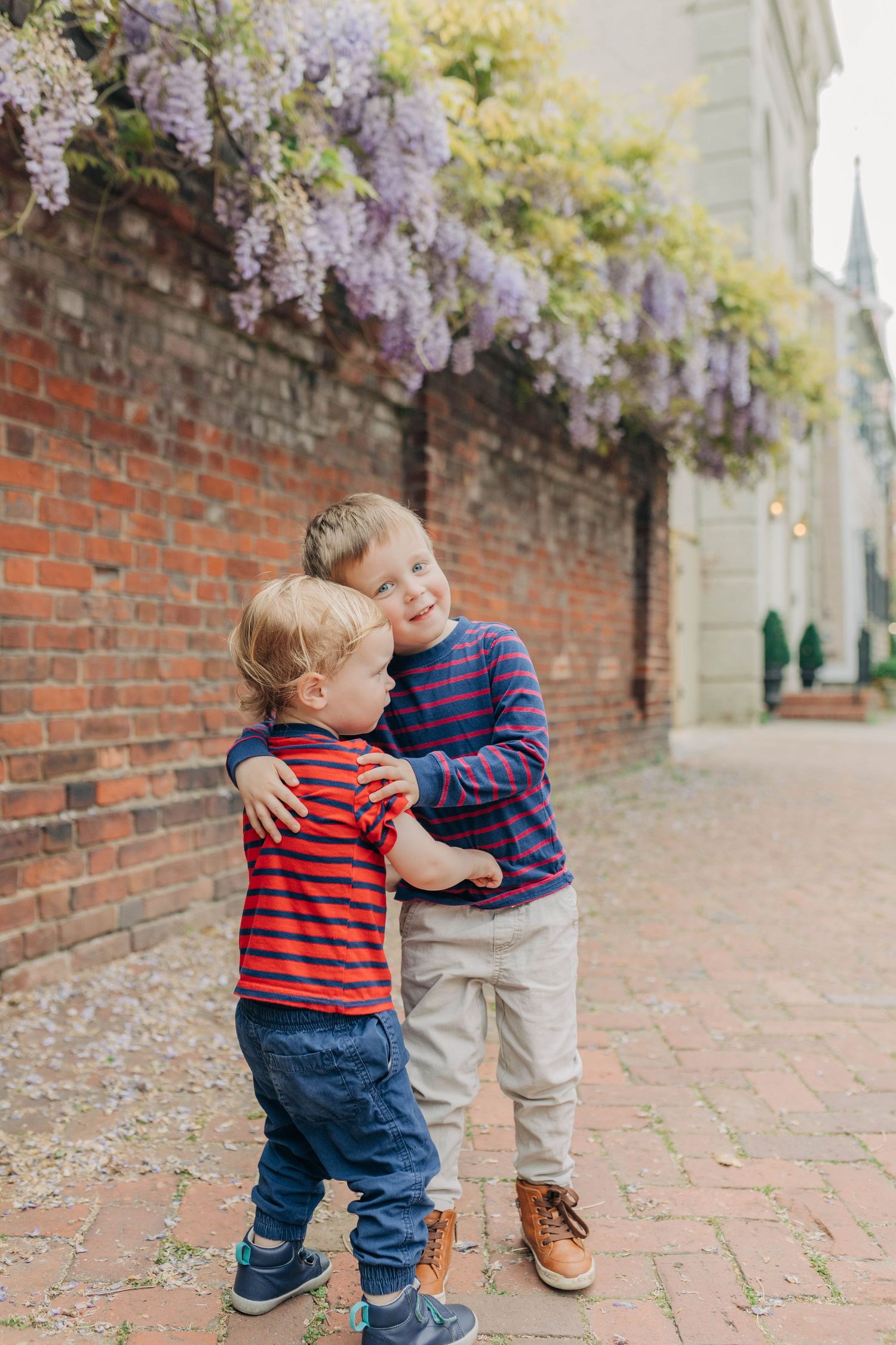 Two children interact playfully on brick sidewalk with purple wisteria flowers overhead.