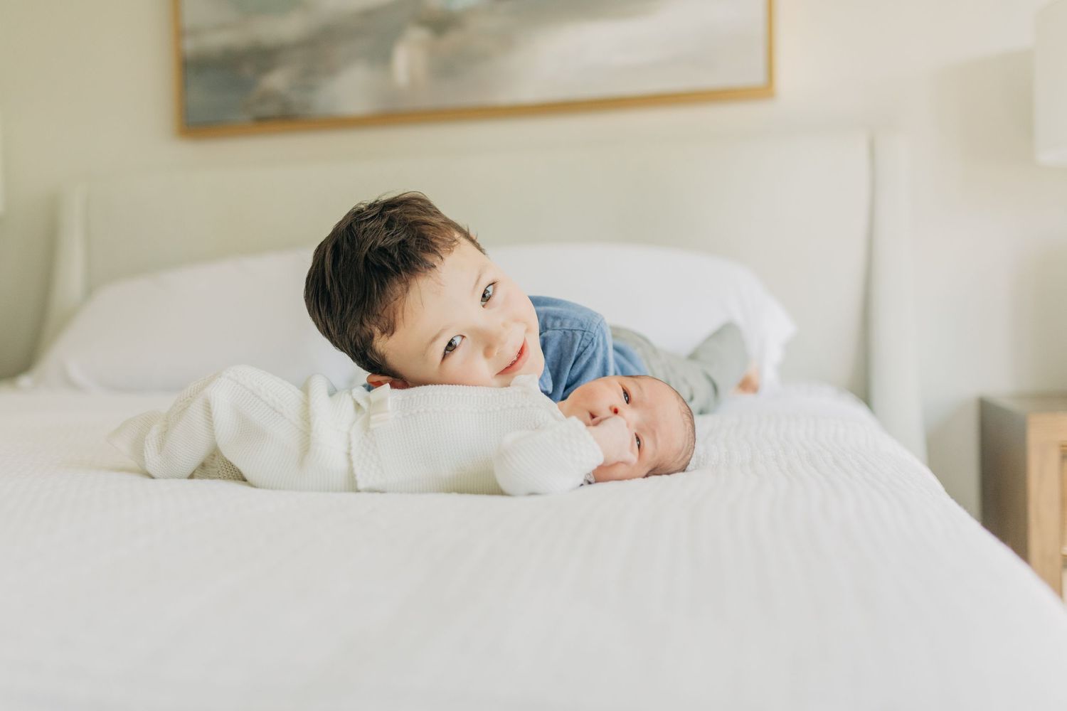 Children share a tender moment together on a white bed with artwork on the wall.