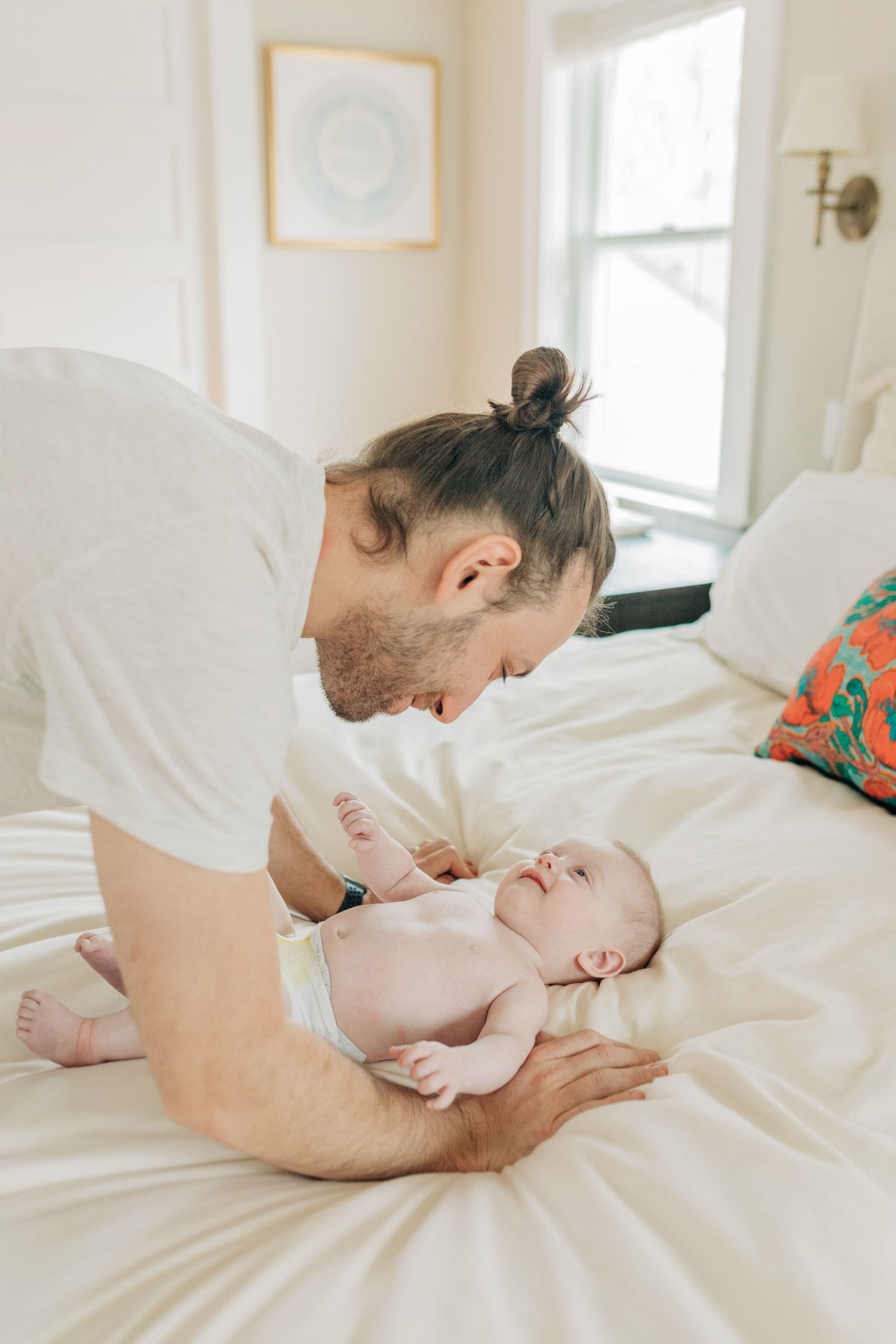 Parent in white shirt leaning over baby on white bedding during intimate moment.