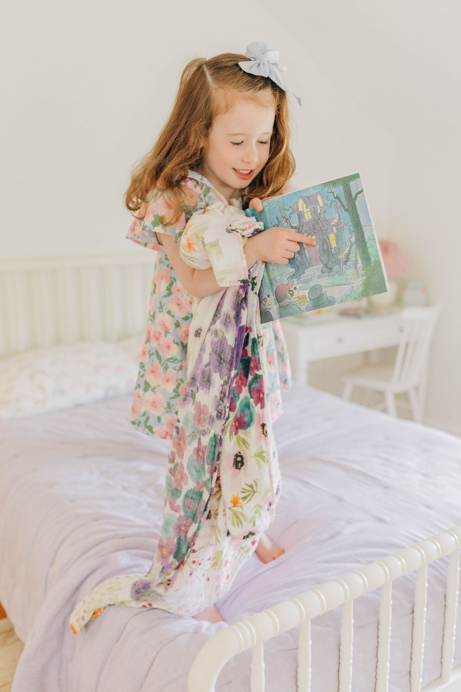 Child in floral dress reading a book while standing on white bedding.