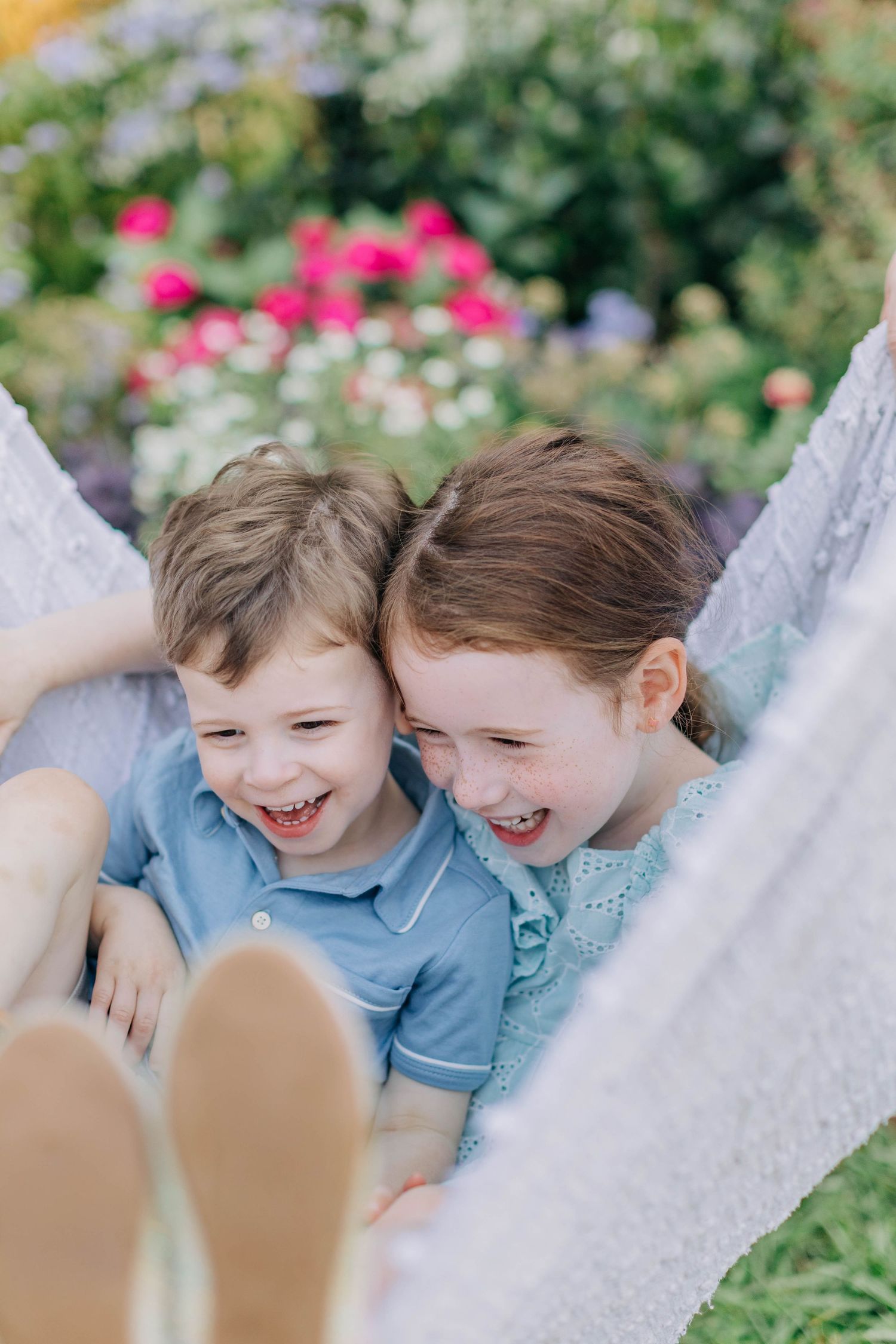 Two children laugh together while relaxing in a hammock surrounded by flowers.