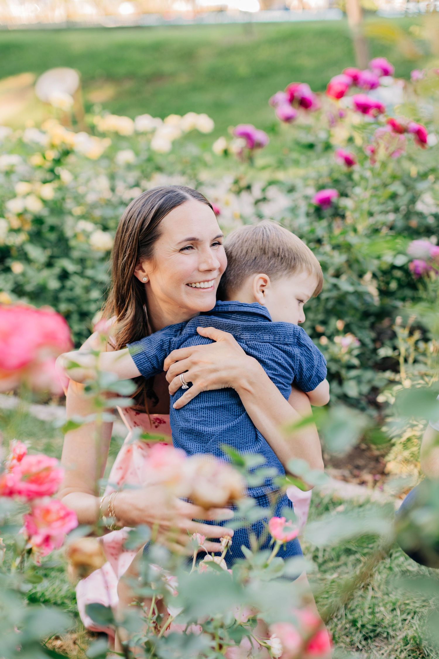 A smiling mother hugs her young son among blooming pink roses in a lush garden.