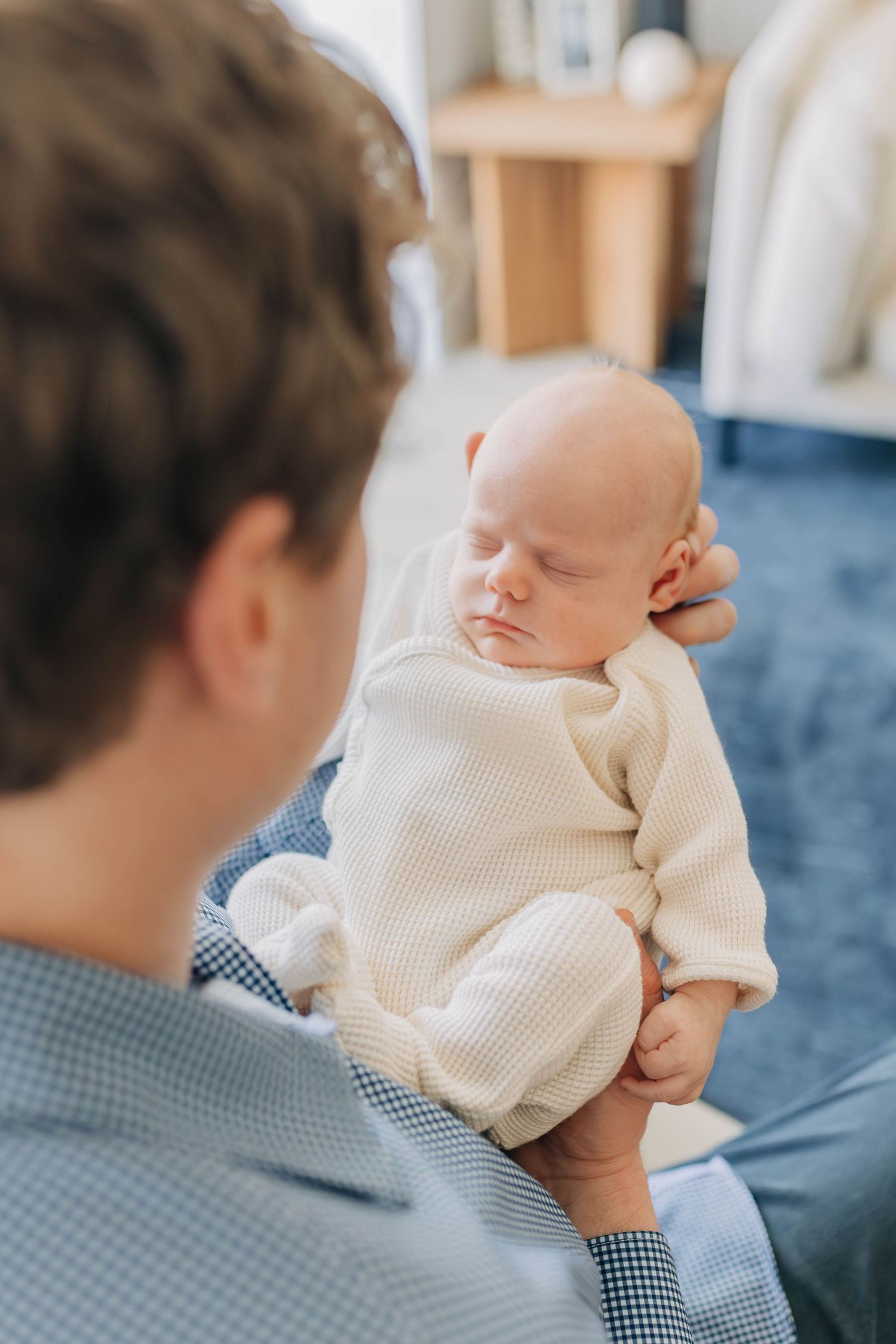 A father gently cradles his sleeping newborn baby dressed in a cream waffle onesie, sharing a tender moment indoors.