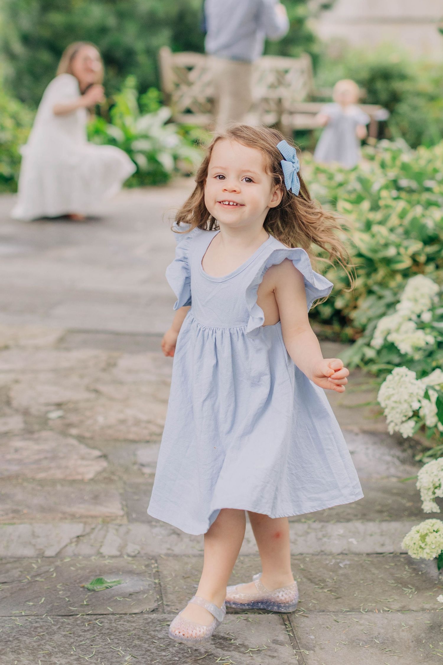 A smiling toddler girl twirls in a light blue ruffle dress in a garden.