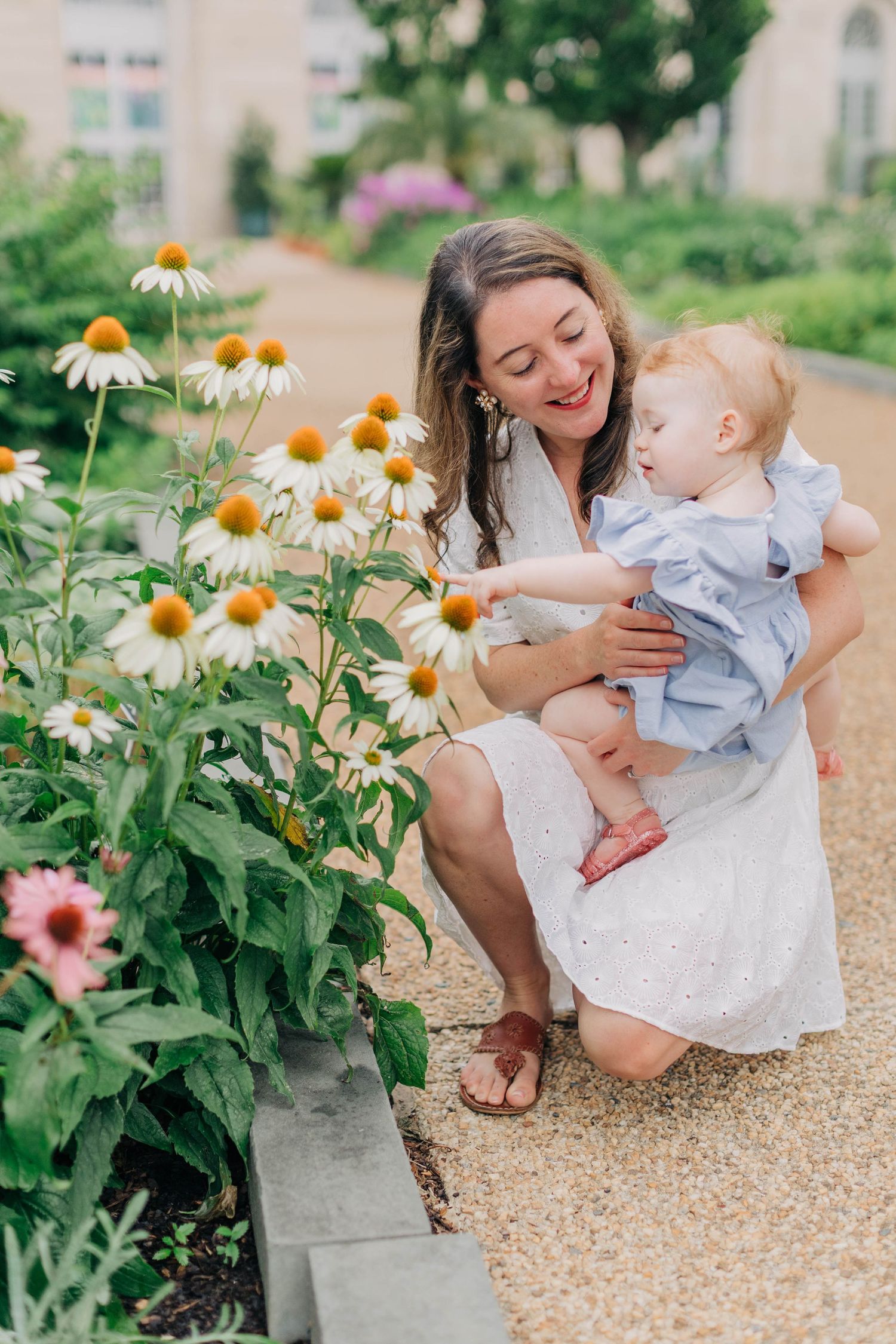 Mother holding baby near white coneflowers in a garden, both smiling as the child reaches to touch the blooms.