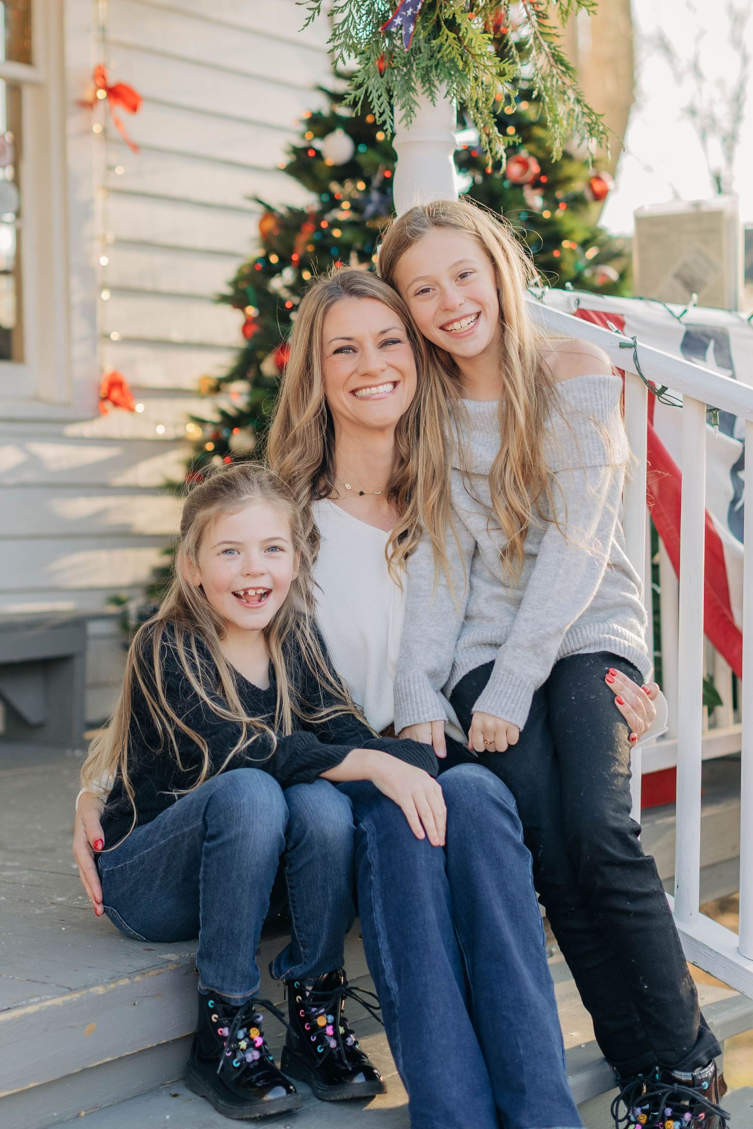 A woman and two girls smile on porch steps in front of a decorated Christmas tree outdoors.