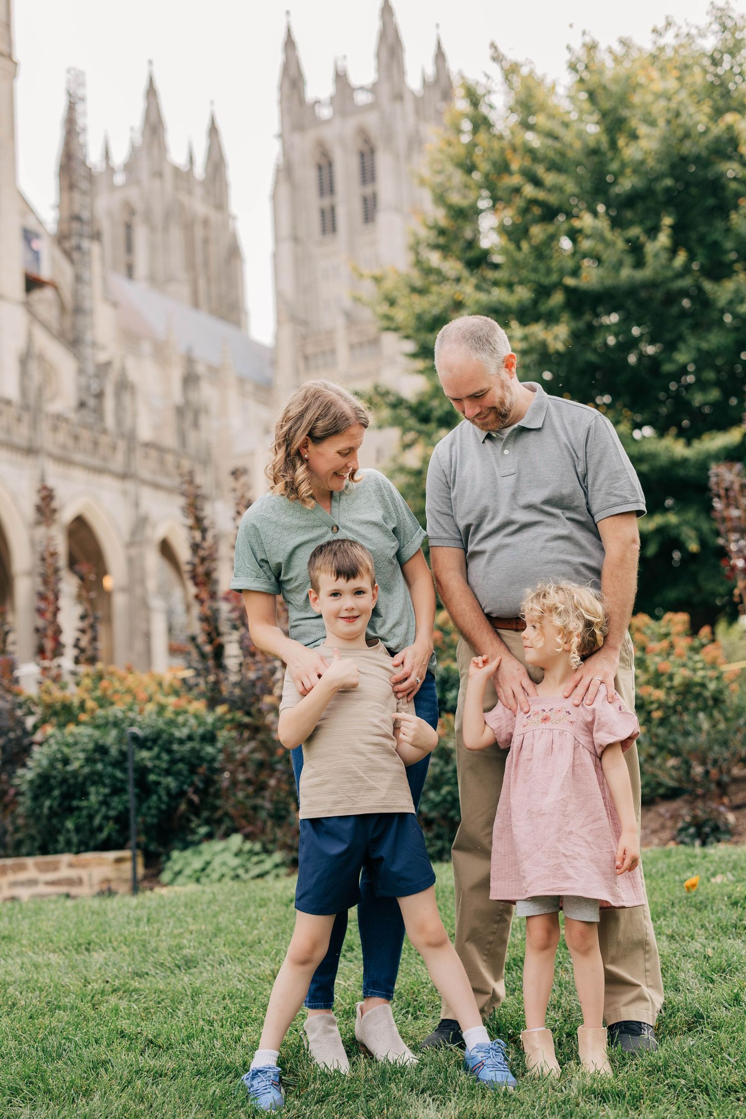 Parents and two young children share a warm moment on a grassy lawn with a Gothic cathedral behind them.