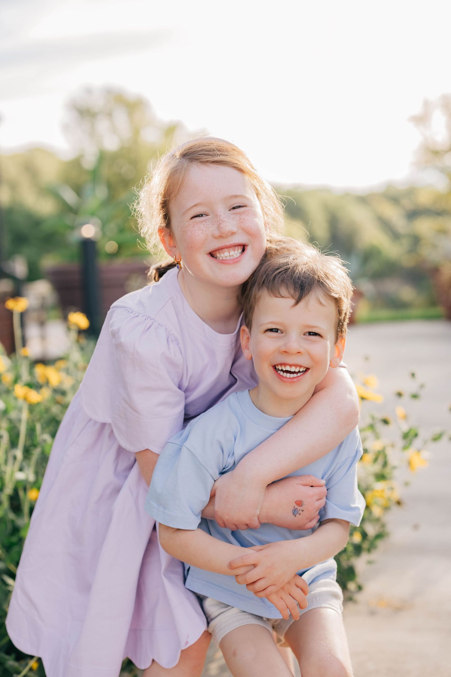 Two happy children, a girl in purple dress and boy in blue shirt, hugging and smiling outdoors with yellow flowers.