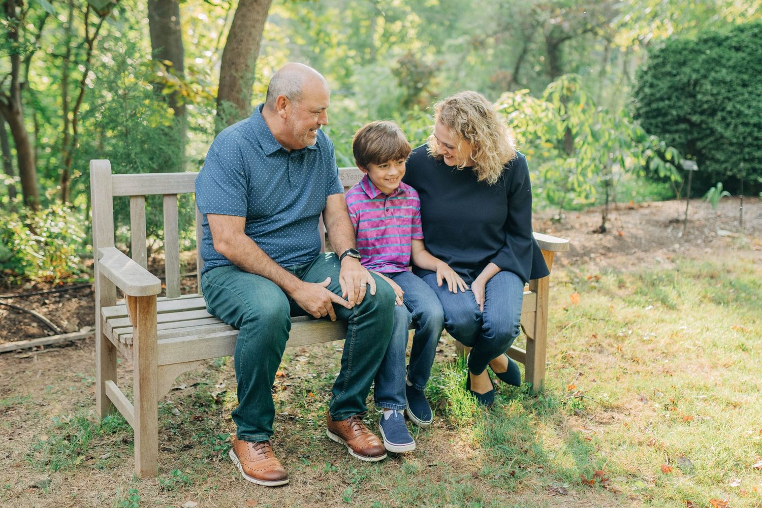 Casual family portraits on wooden bench with lush garden and trees in background.