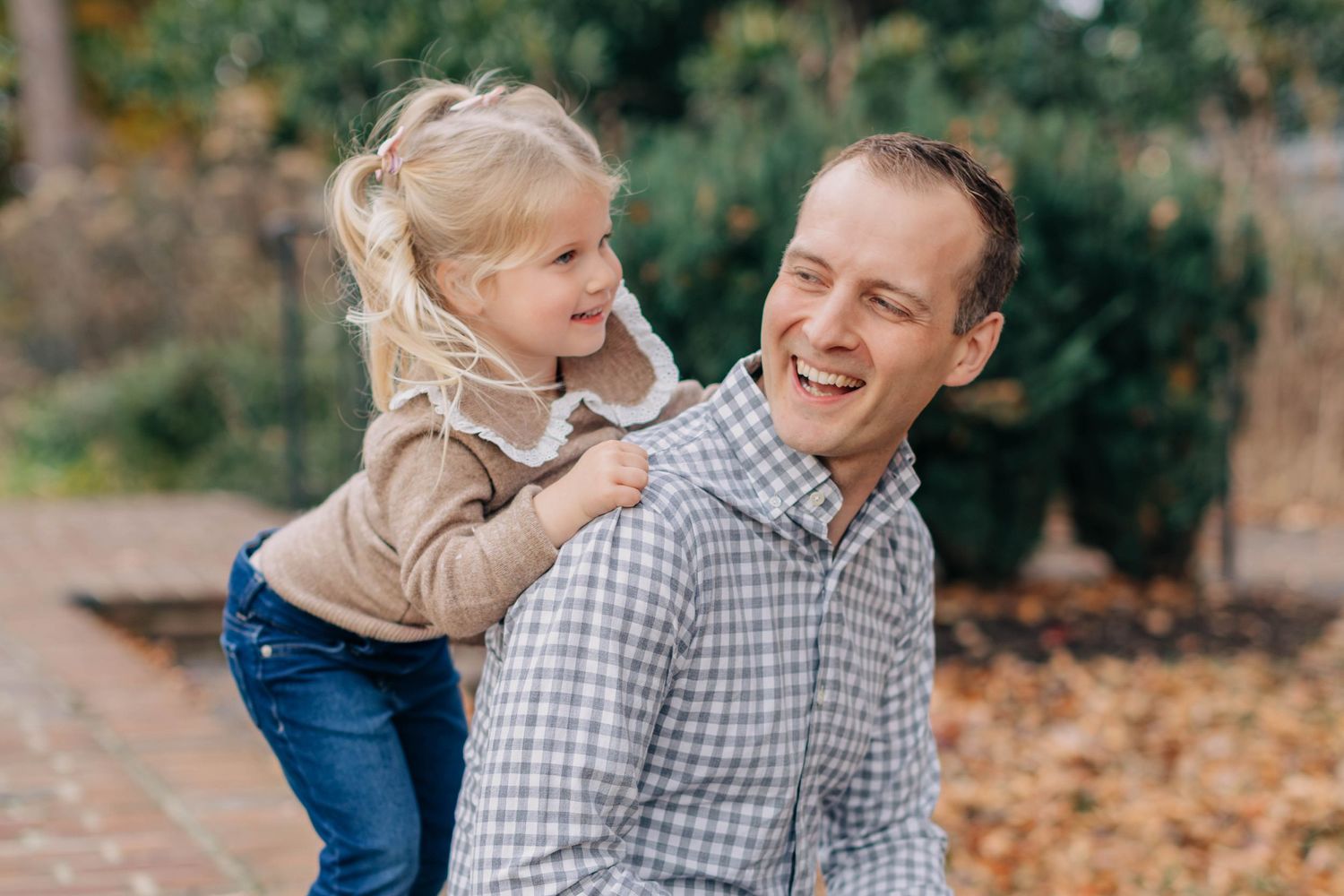 A smiling man carries a laughing little girl on his back outdoors in a park during autumn.