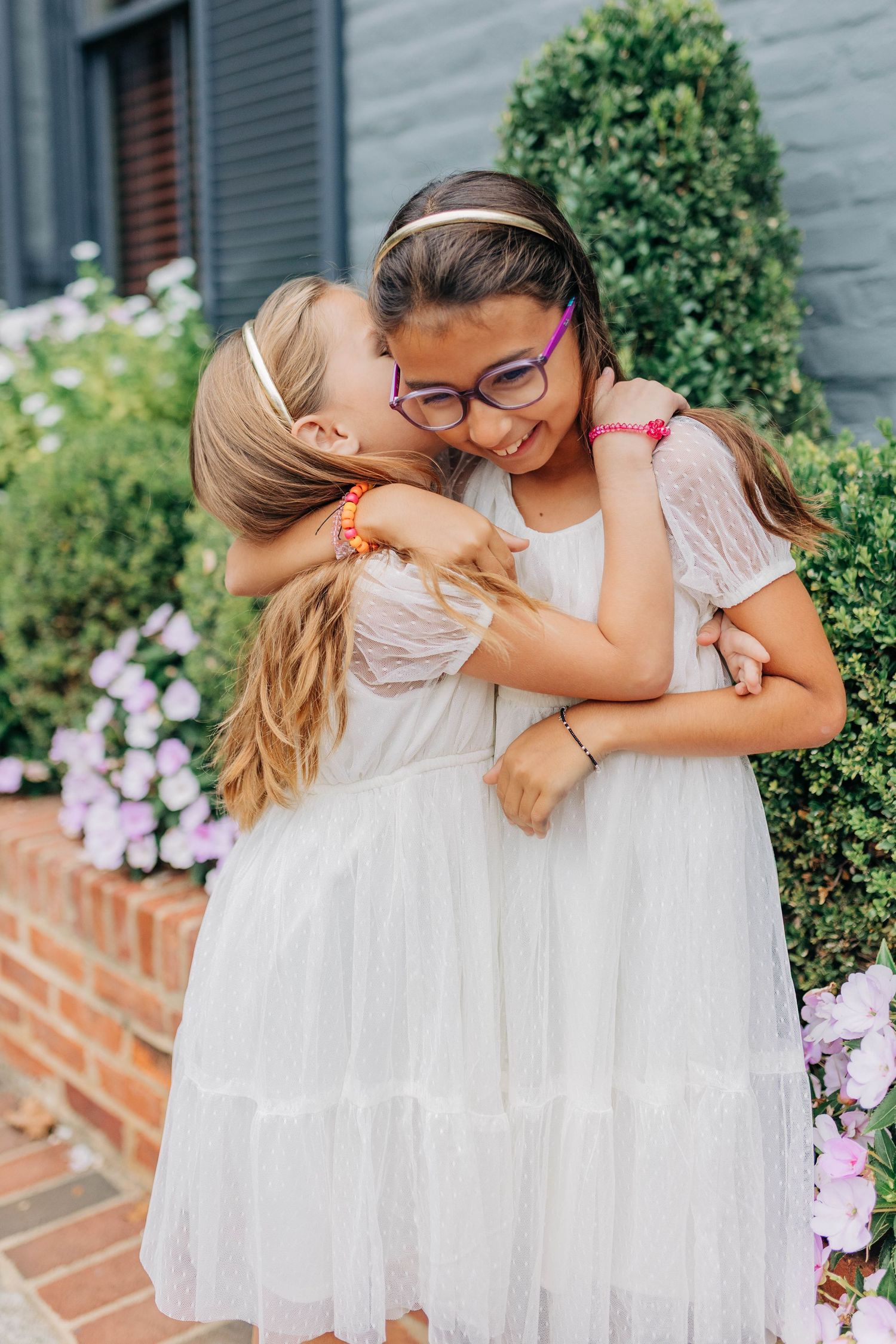 Two young girls in white dresses hugging and sharing a kiss outdoors near blooming flowers and green shrubs.