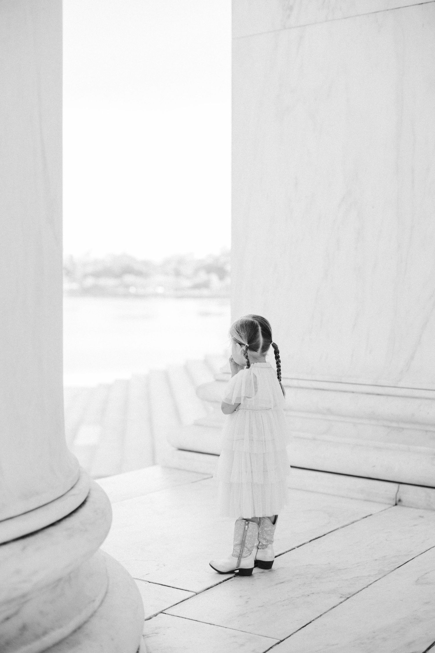 A young girl in a white dress and braids stands between marble columns at the Jefferson Memorial in Washington D.C.
