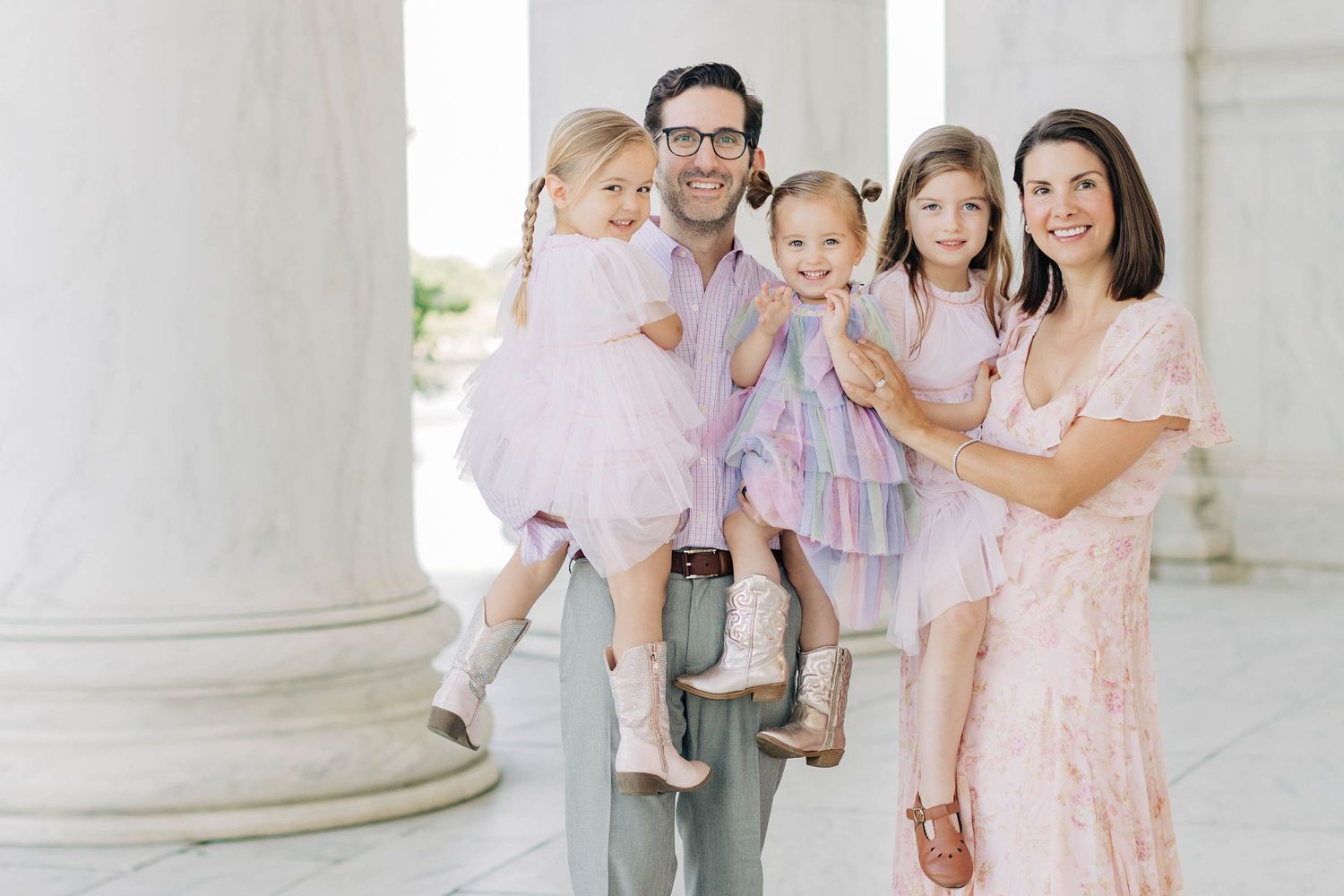 Happy family of five posing outdoors near white columns, parents holding three young girls in coordinating pink outfits.