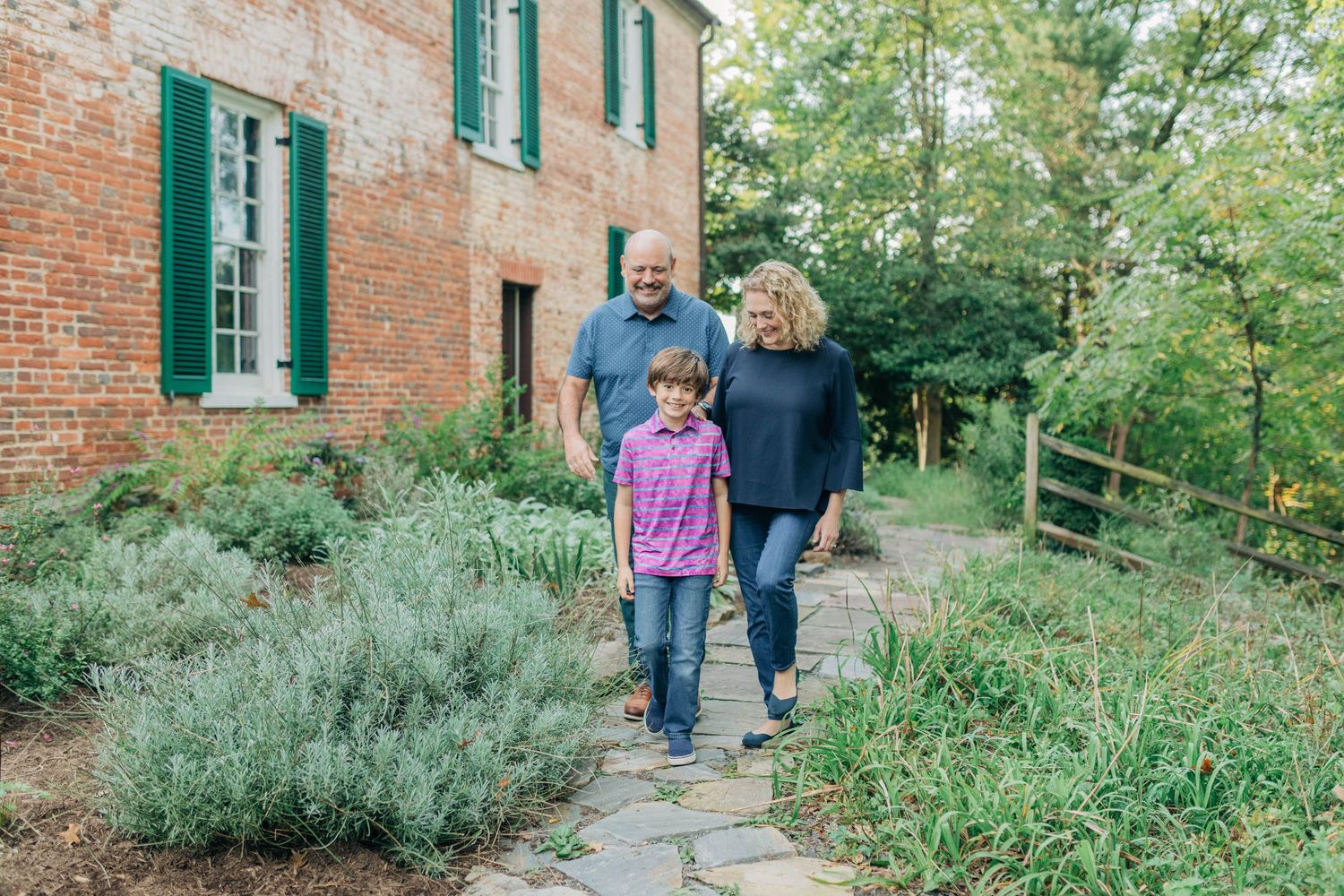 Family walks together on stone path alongside brick building with green shutters and garden beds.