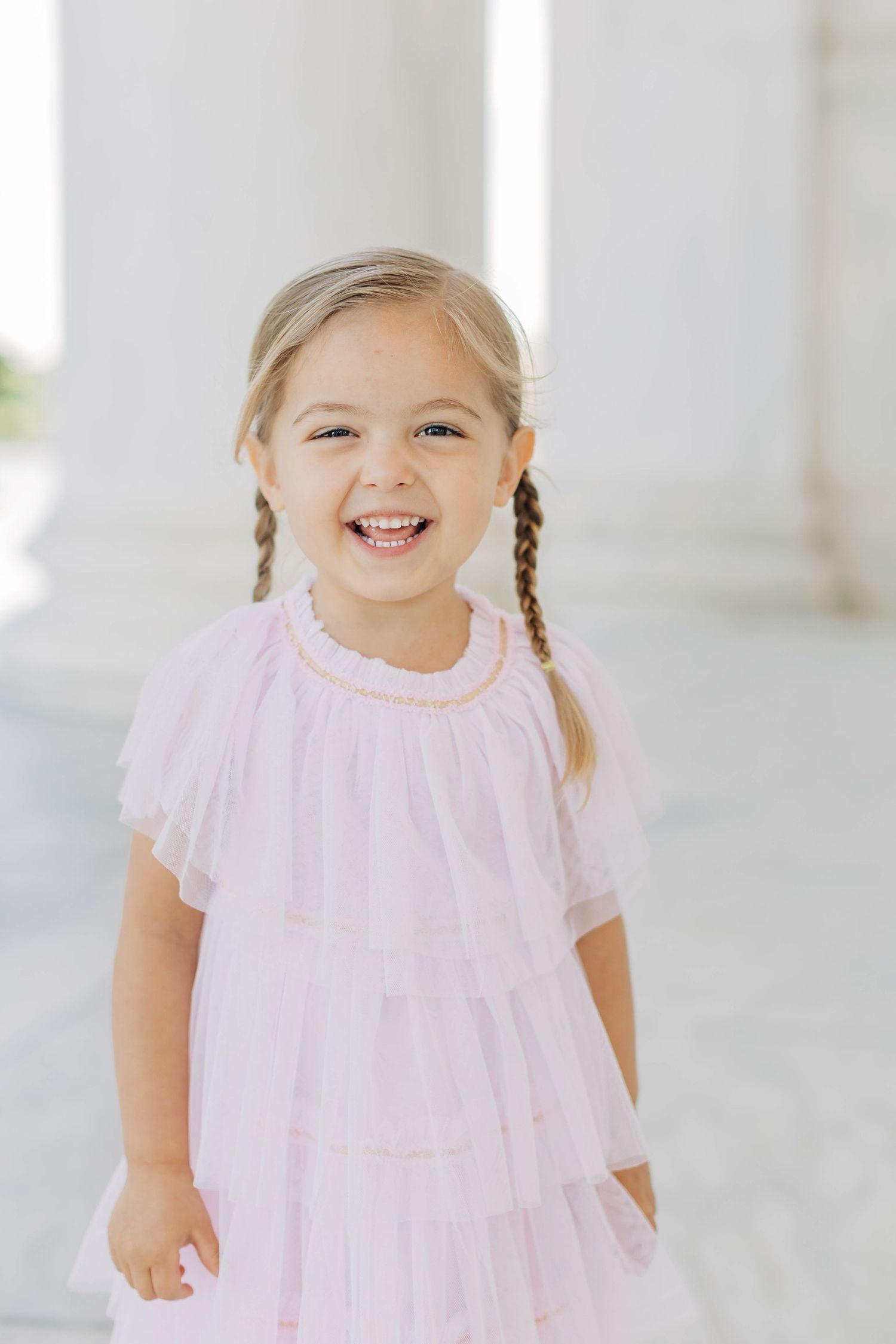 Smiling young girl with pigtail braids wearing a pink tiered tulle dress near white columns.