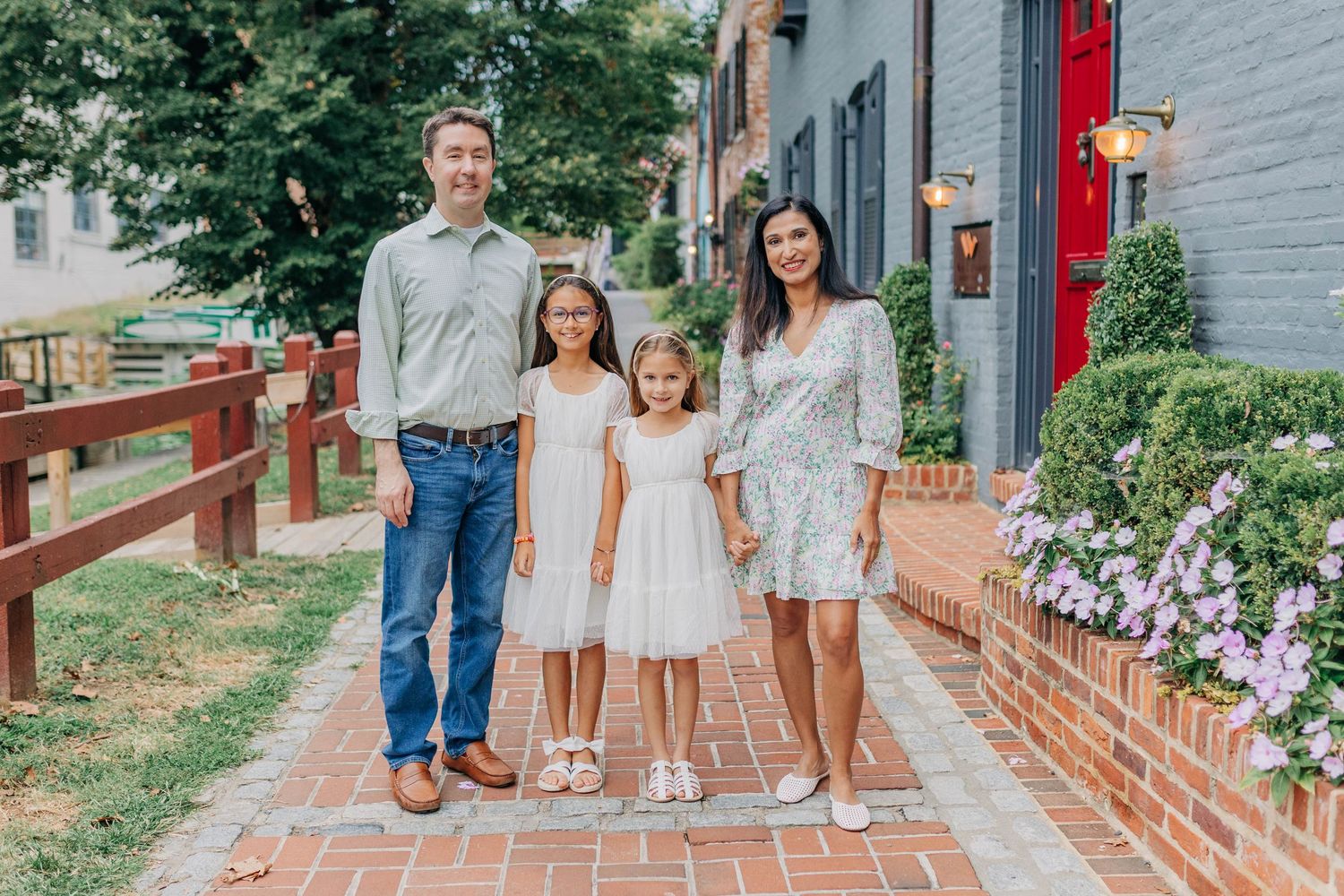 Family of four posing on a brick pathway beside a gray building with colorful flowers.